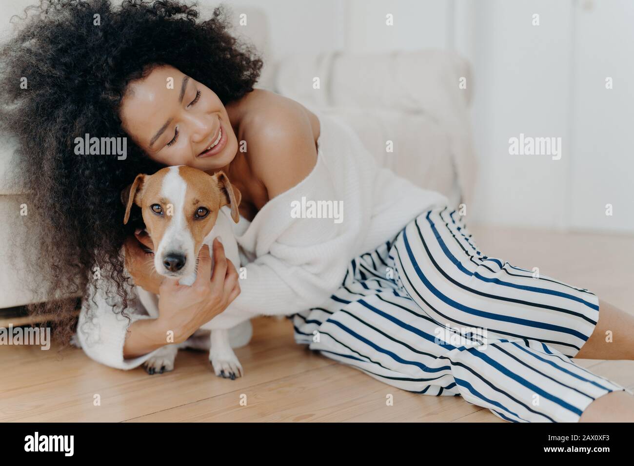 Affectueuse jeune femme hople chien avec amour et soin, garde les yeux fermés du plaisir, sourires doucement, a une peau sombre saine, pose sur le sol, petting A. Banque D'Images