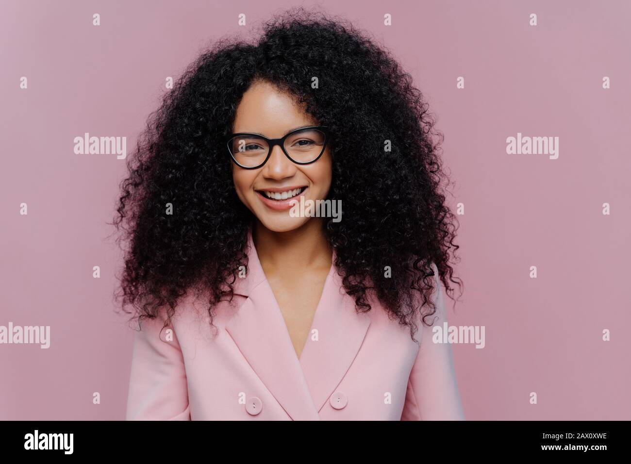Gros plan portrait de heureuse femme aux cheveux bouclés a le sourire doux sur le visage, peau sombre saine cheveux bushy, porte des vêtements élégants, isolé sur le dos violet Banque D'Images