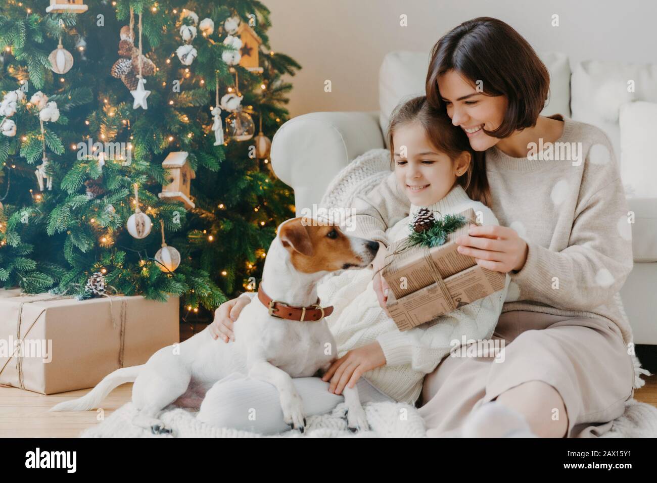 Une jeune femme heureuse embrasse sa petite fille, présente Noël, attend pour des vacances d'hiver, jouer avec le chiot pedigree, poser au sol près de la décoration Banque D'Images