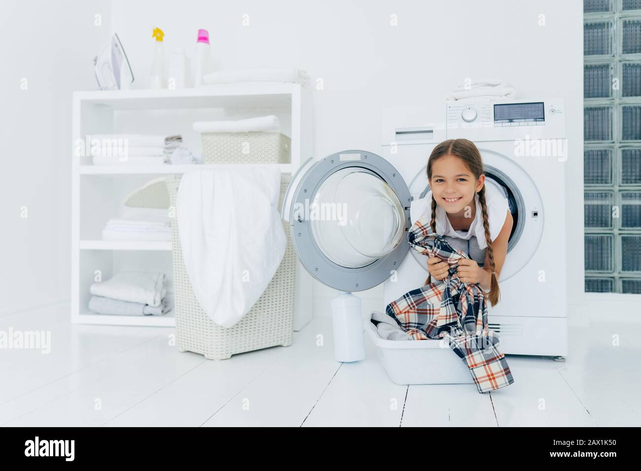 Enfant positif avec des bâtons de queue de porc hors de la tête de la machine à laver, s'amuse et tient la chemise, se prépare pour le lavage, sourires doucement, passe du temps libre à lau Banque D'Images