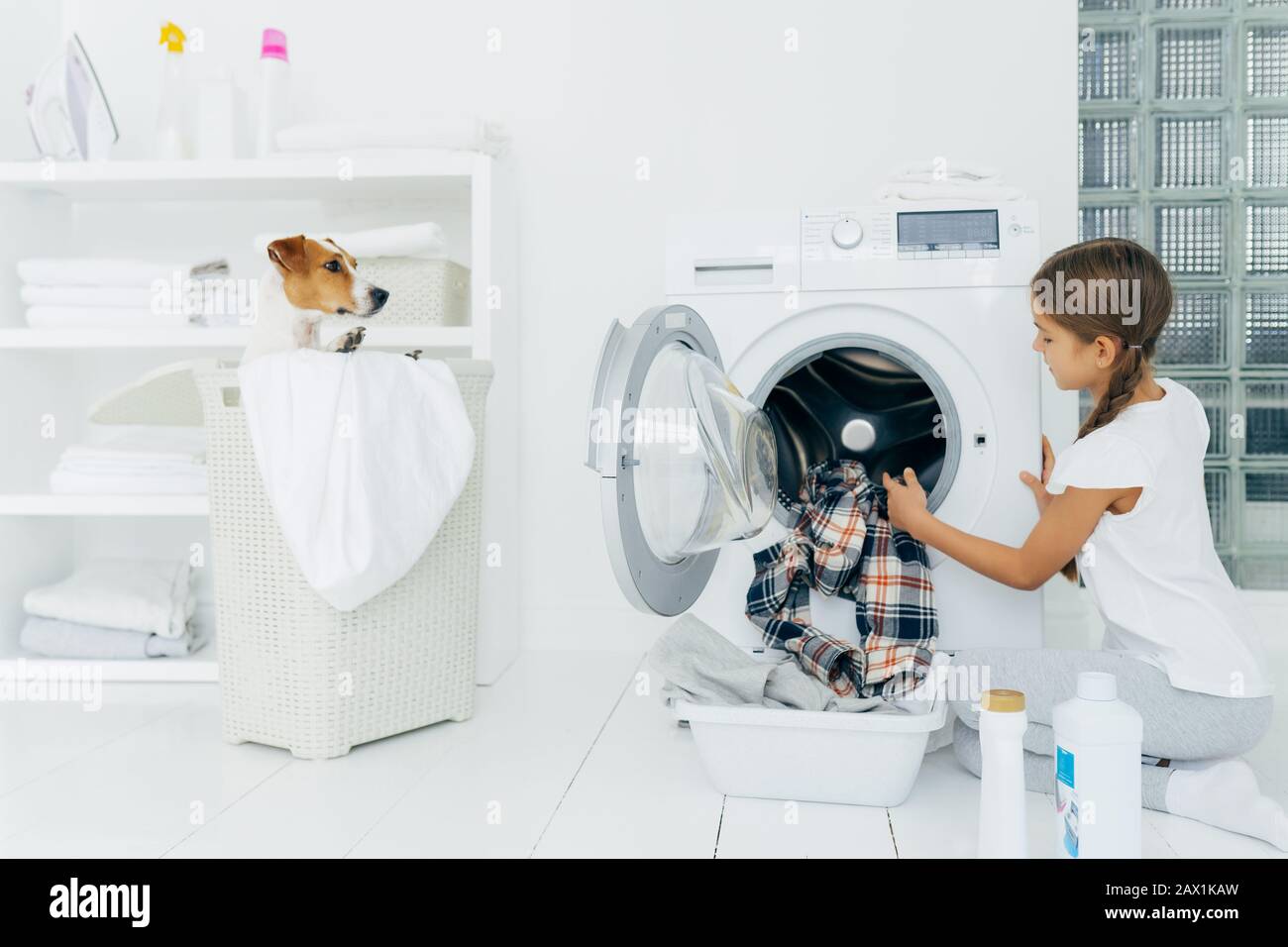L'enfant occupé fait le travail de blanchisserie, vide la machine à laver, les vêtements nettoyés dans le bassin utilise des détergents, petit chien pedigree dans le panier. Moderne ménage dévic Banque D'Images