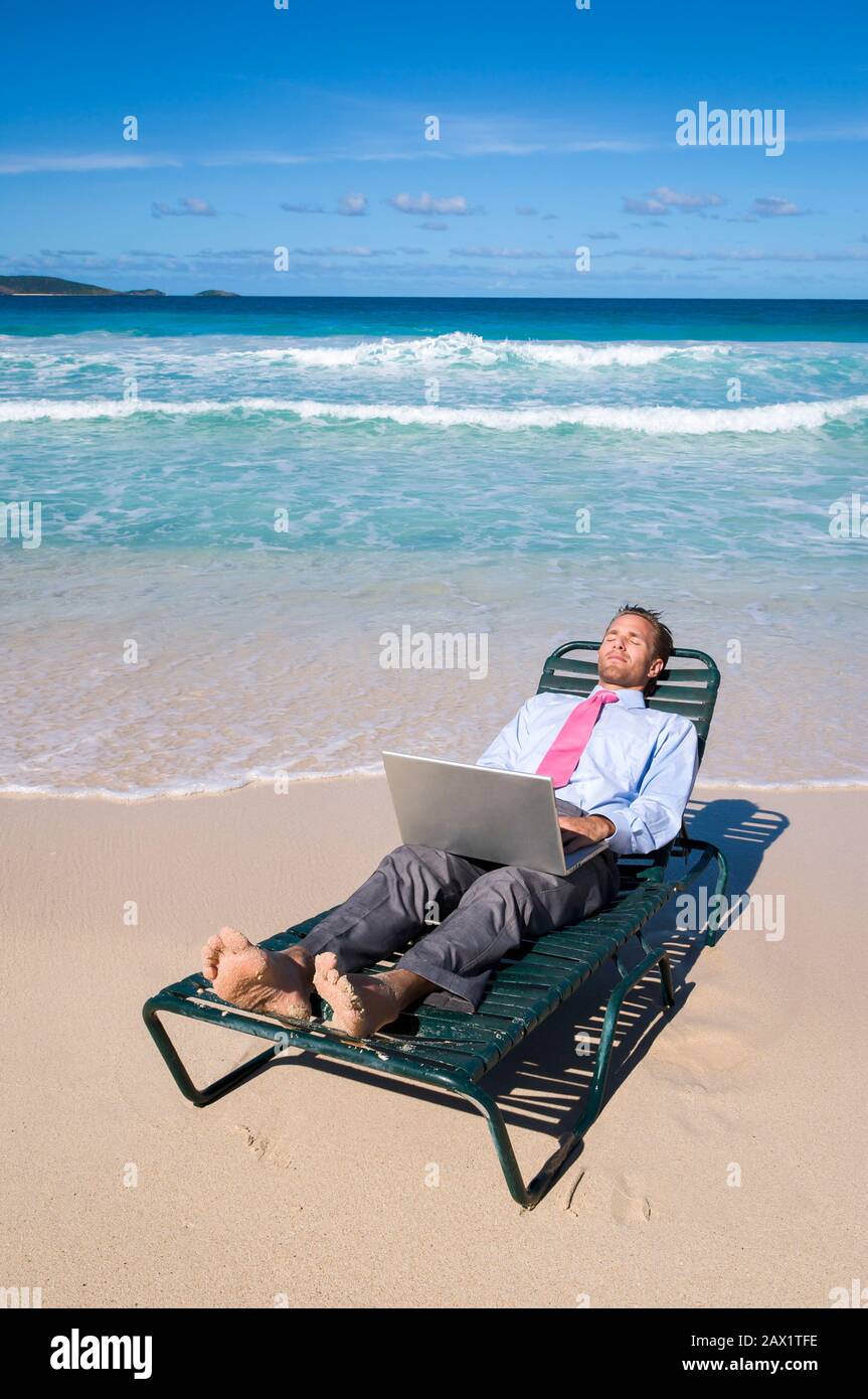 Homme d'affaires pieds nus travaillant sur son ordinateur portable sur une chaise de plage se reposant sur la rive d'une plage tropicale vide Banque D'Images