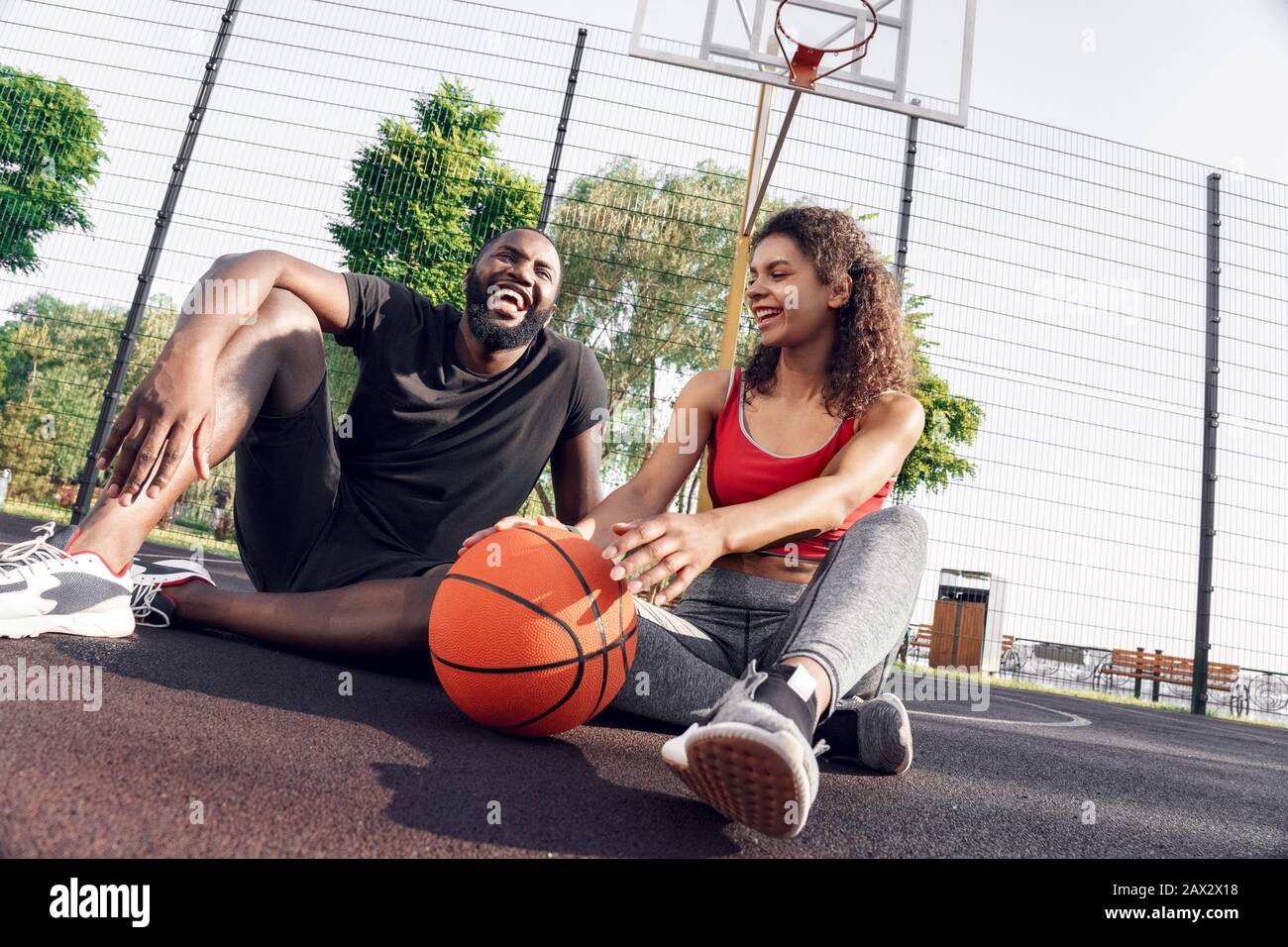 Activité En Plein Air. Couple africain assis avec ballon sur le terrain de basket-ball en riant la vue de fond heureuse Banque D'Images