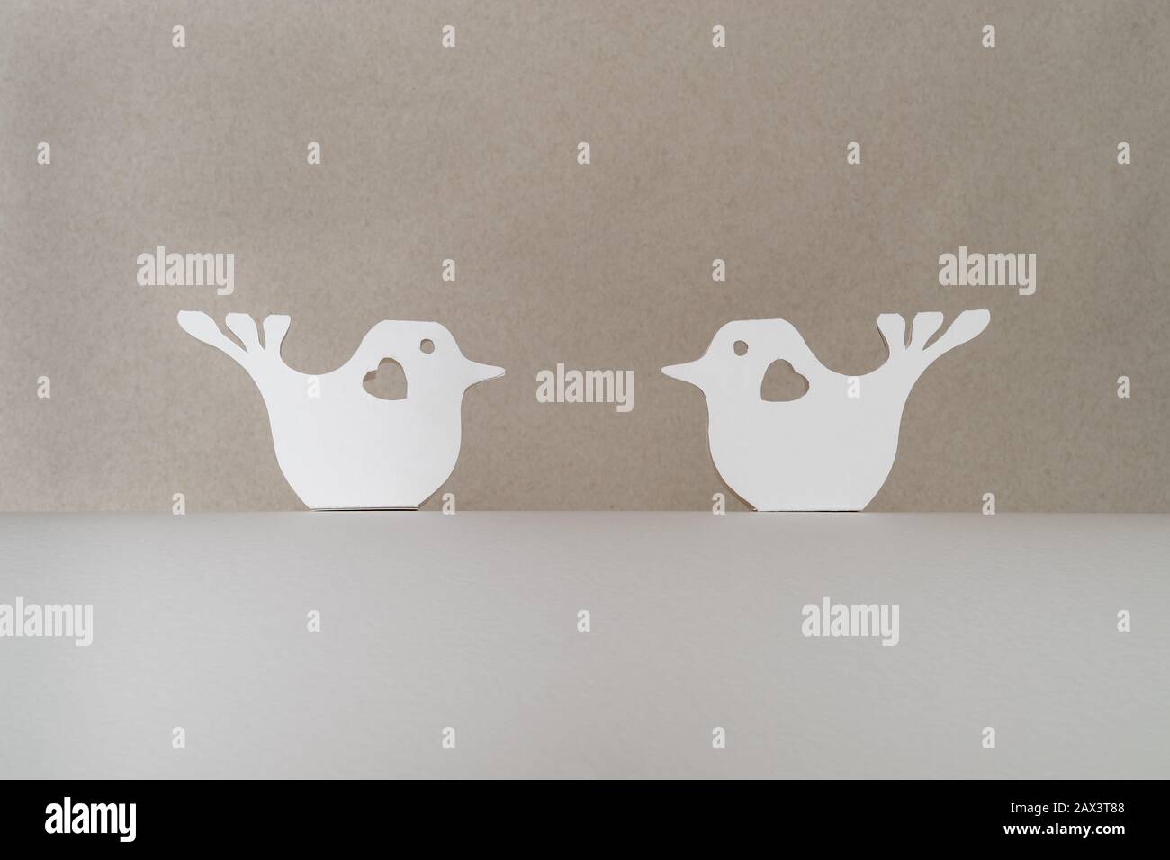 Deux oiseaux blancs coupés en papier avec des coeurs assis l'un contre l'autre sur une base de carton blanc sur un fond beige. Saint Valentin et concept d'amour Banque D'Images