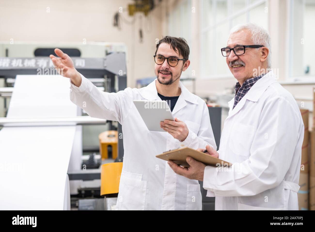Un expert souriant de qualité barbu dans les verres gestant la main et utilisant la tablette tout en discutant de la productivité de l'équipement de maison d'impression avec un collègue Banque D'Images