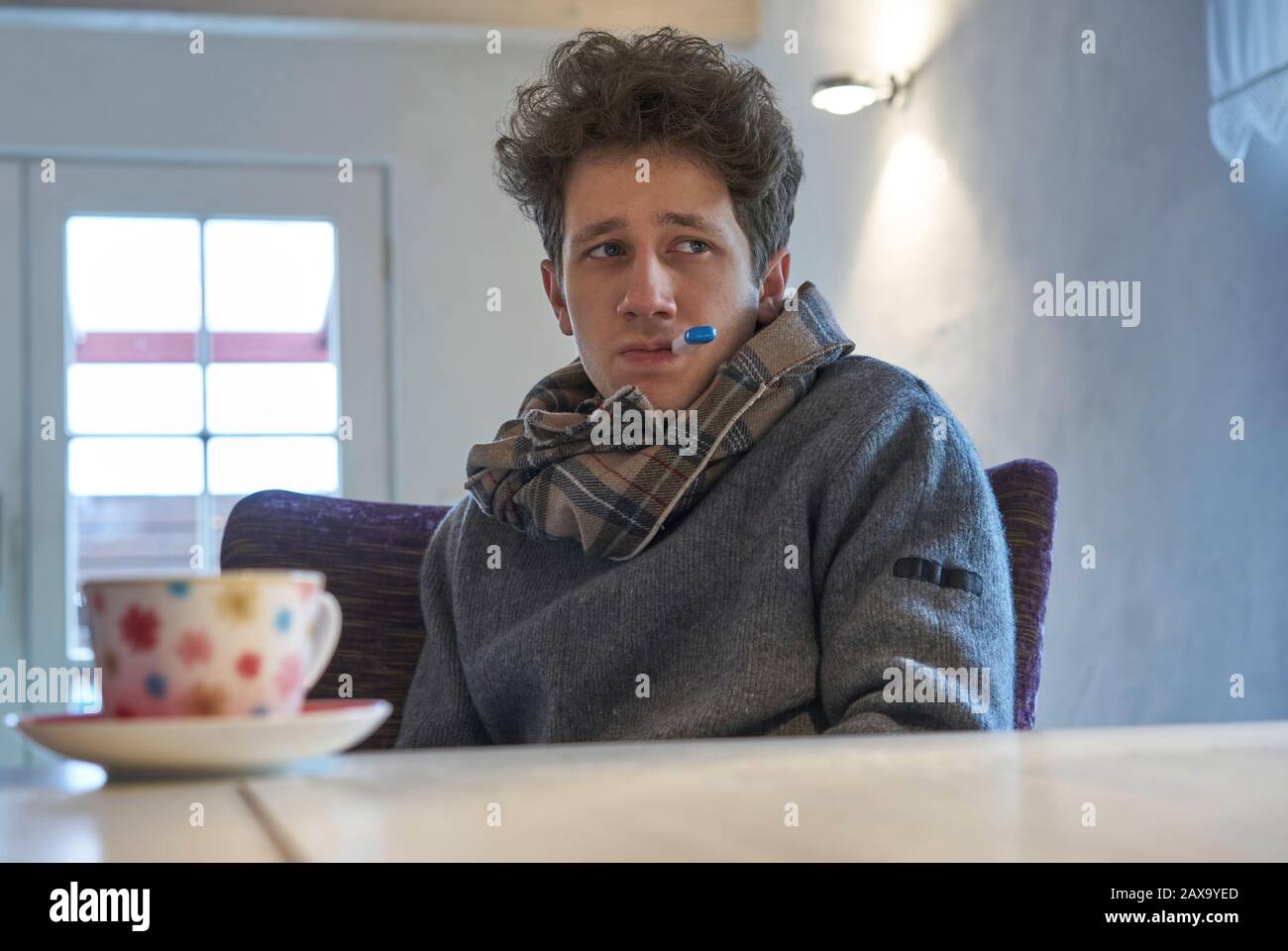 le nuage de jeune homme se trouve à la table à la maison avec un foulard autour de son cou et a un thermomètre dans sa bouche, il semble comme s'il a un froid Banque D'Images