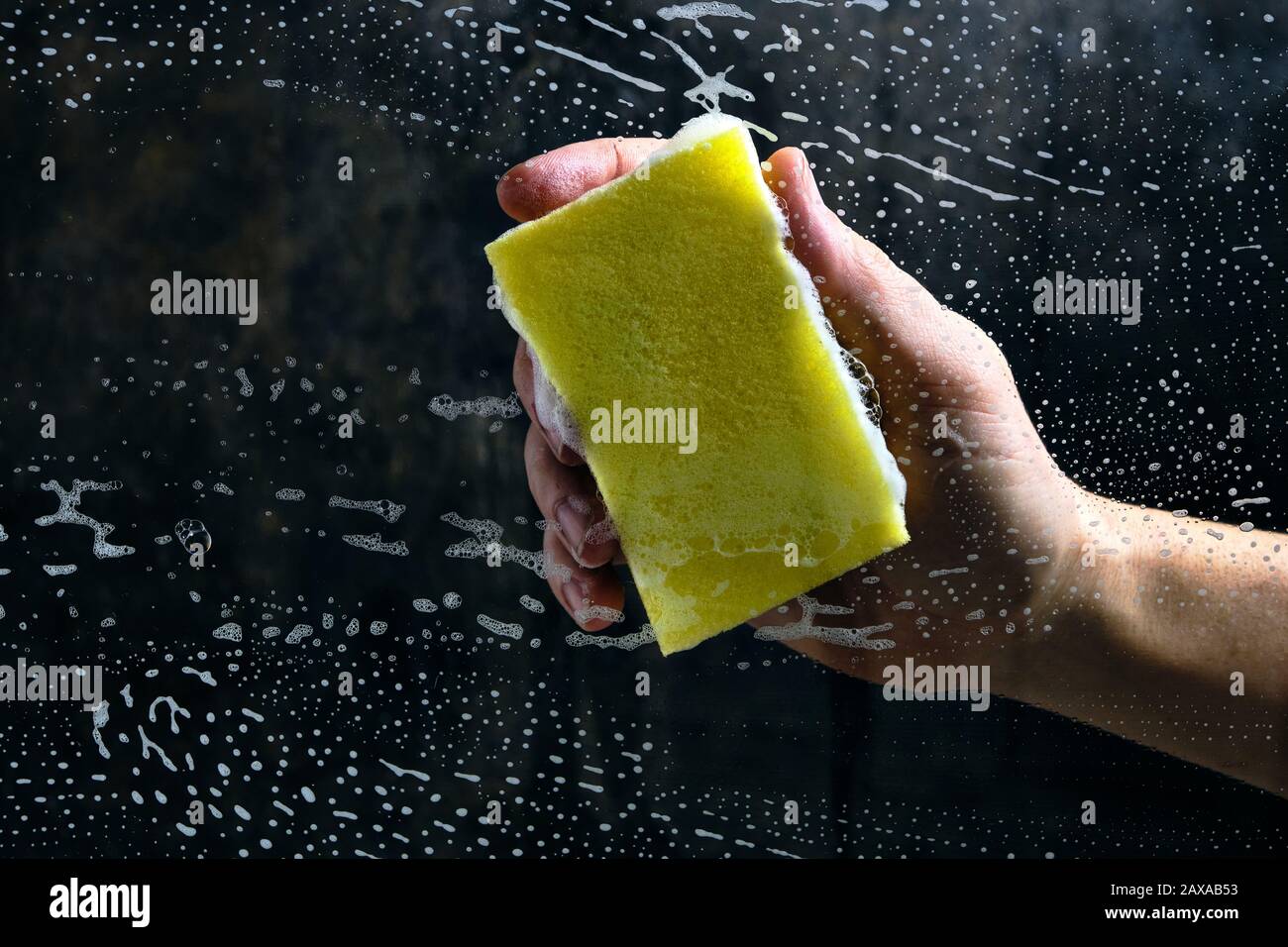 La main de l'homme lave le verre recouvert de mousse, éponge jaune sur fond sombre Banque D'Images