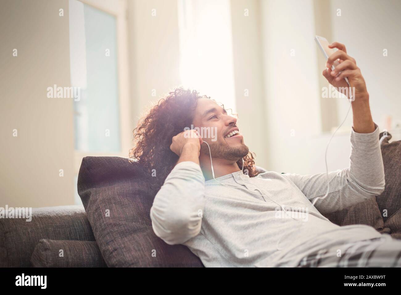 Un jeune homme souriant se détendant, écoutant de la musique avec un smartphone et un casque sur un canapé Banque D'Images