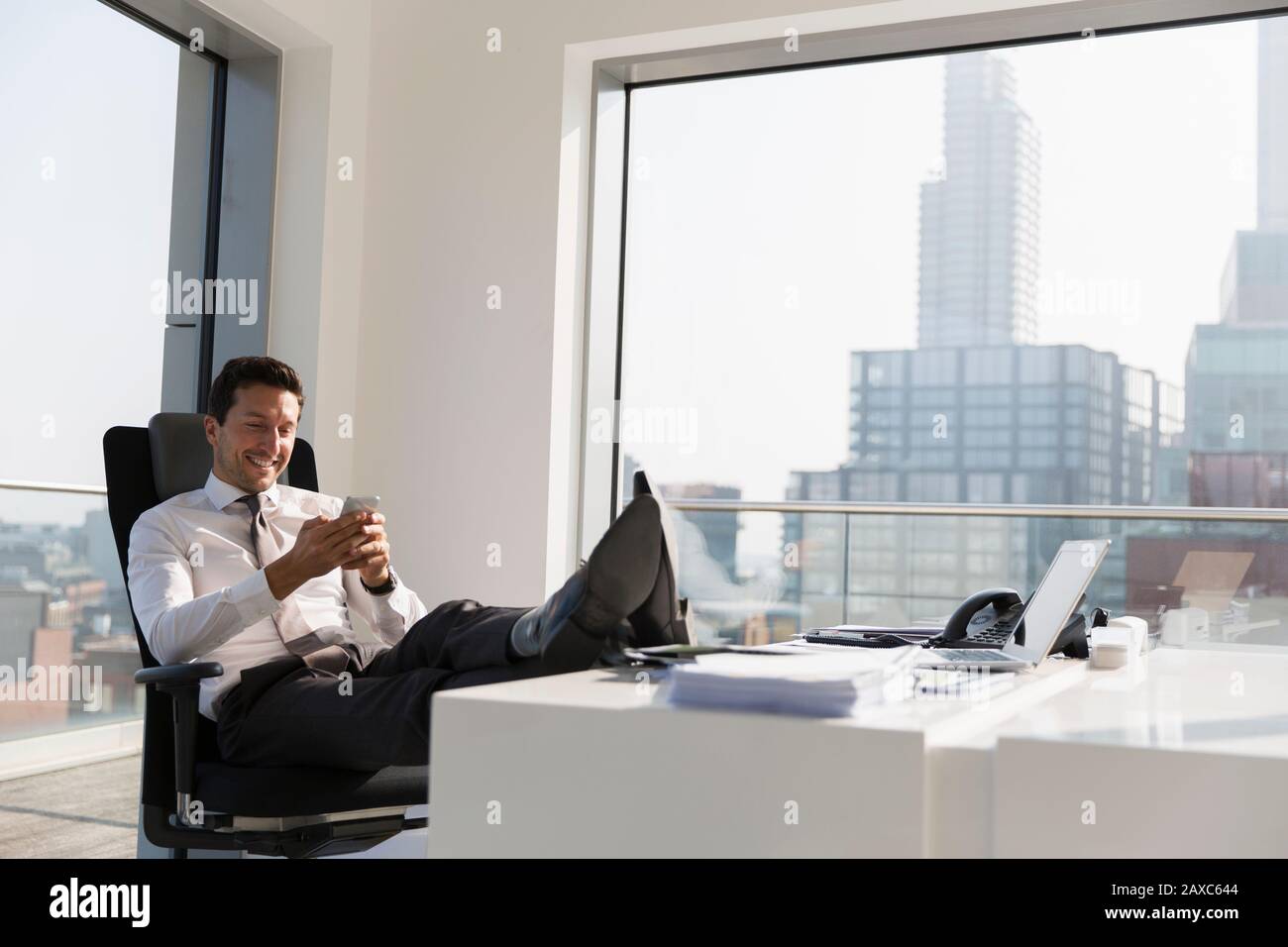 Homme d'affaires souriant utilisant un smartphone avec des pieds sur le bureau dans un bureau moderne, ensoleillé, urbain Banque D'Images