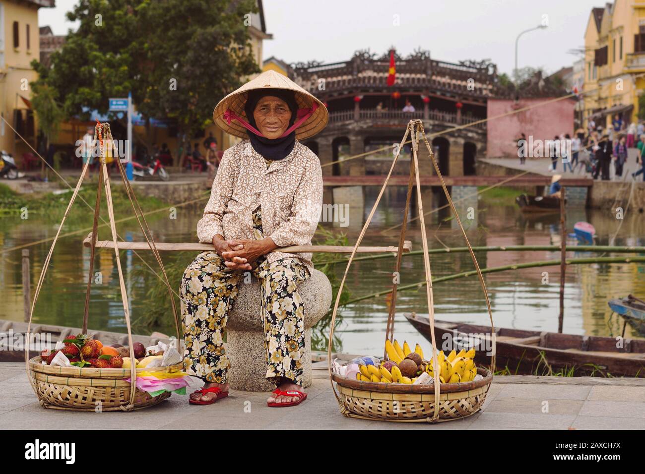Vieille dame vietnamienne assise avec des paniers de fruits devant le pont Couvert japonais à Hoi An, au Vietnam. Banque D'Images