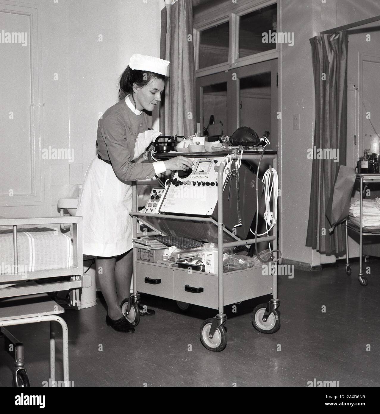Années 1960, historique, infirmière dans un service hospitalier avec un chariot avec un défibrillateur DC, un dispositif électronique qui est utilisé pour ranimer le cœur d'un patient après une urgence cardiaque, South London, Angleterre, Royaume-Uni. Banque D'Images
