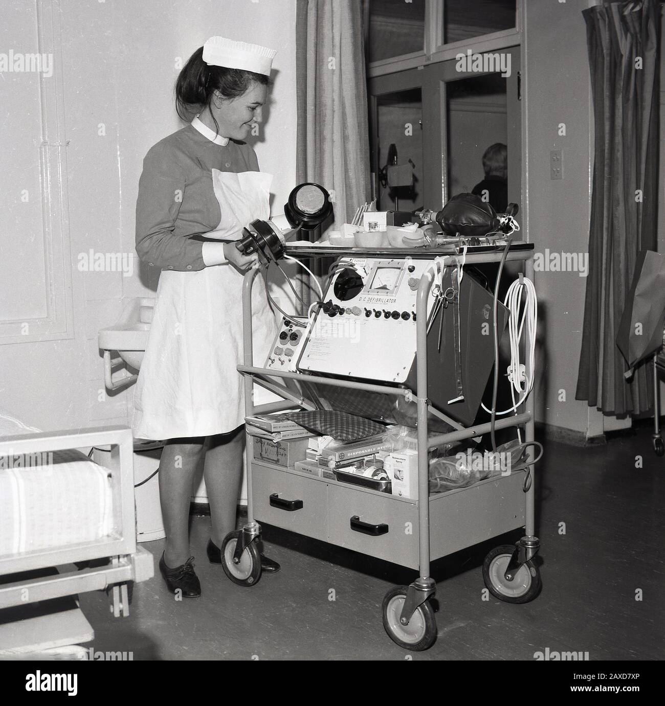 Années 1960, historique, infirmière dans un service hospitalier avec un chariot avec un défibrillateur DC, un dispositif électronique qui est utilisé pour ranimer le cœur d'un patient après une urgence cardiaque, South London, Angleterre, Royaume-Uni. Banque D'Images