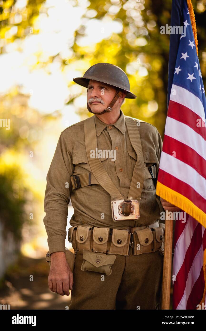 Le réacteur masculin mature portant un casque de combat en acier de la guerre mondiale et un uniforme et tenant un drapeau américain pose un portrait alors qu'il se tient sur le trottoir d'une rue de banlieue. Banque D'Images