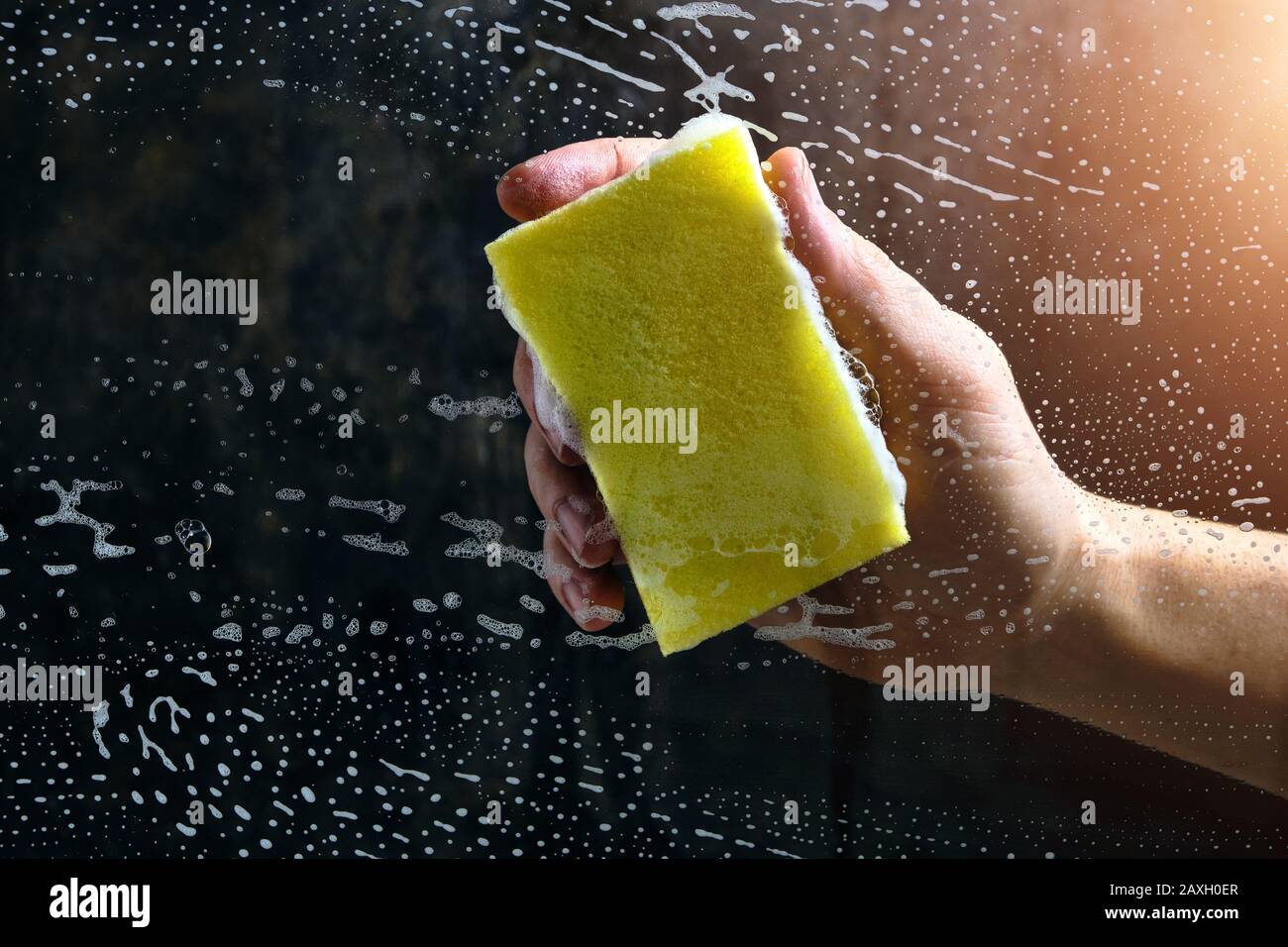 La main de l'homme lave le verre recouvert de mousse, éponge jaune sur fond sombre Banque D'Images