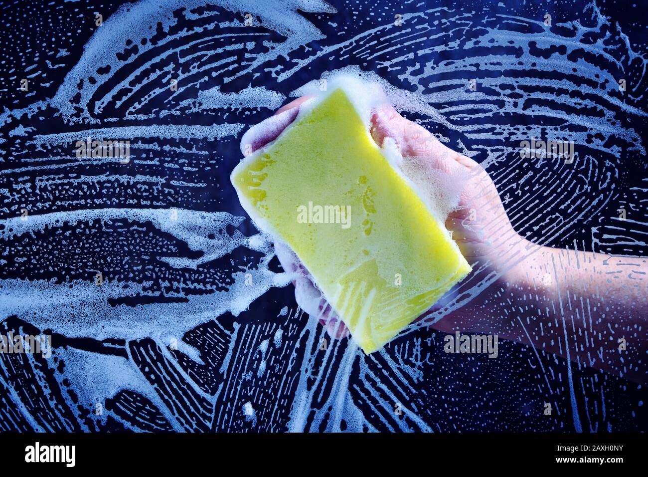 La main de l'homme lave le verre recouvert de mousse, éponge jaune sur fond sombre Banque D'Images