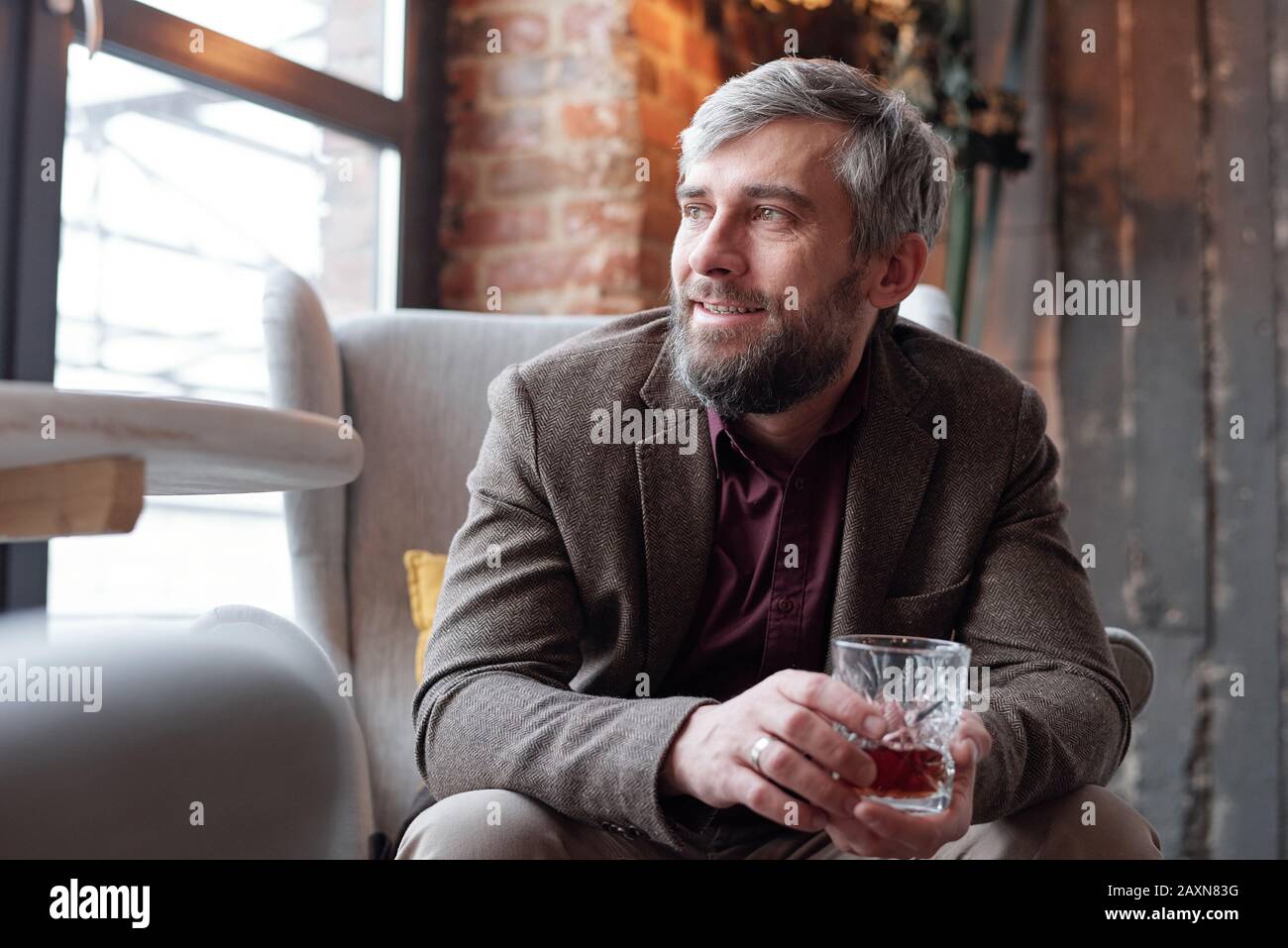Homme à la barbe de rêve d'âge moyen dans une veste marron assise dans un fauteuil et buvant de l'alcool Banque D'Images