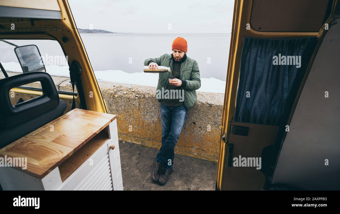 Jeune homme ayant une boisson chaude de thermos devant une fourgonnette de camping-cars donnant sur un lac en arrière-plan Banque D'Images