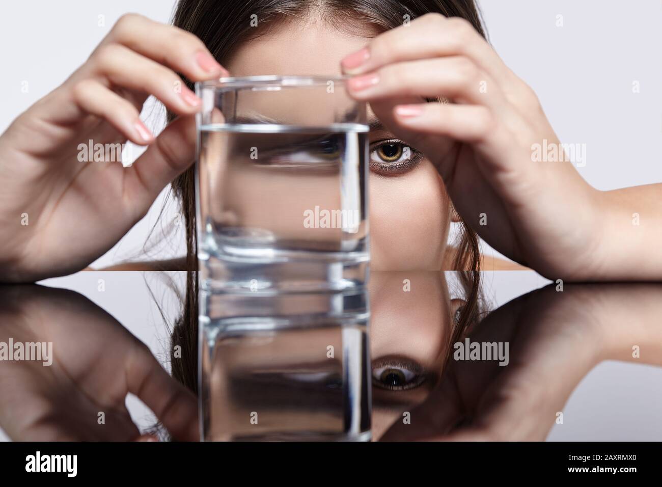 Fille cache son visage derrière un verre avec de l'eau. Portrait de distorsion optique de la jeune femme à la table miroir. Femme sur fond gris. Banque D'Images
