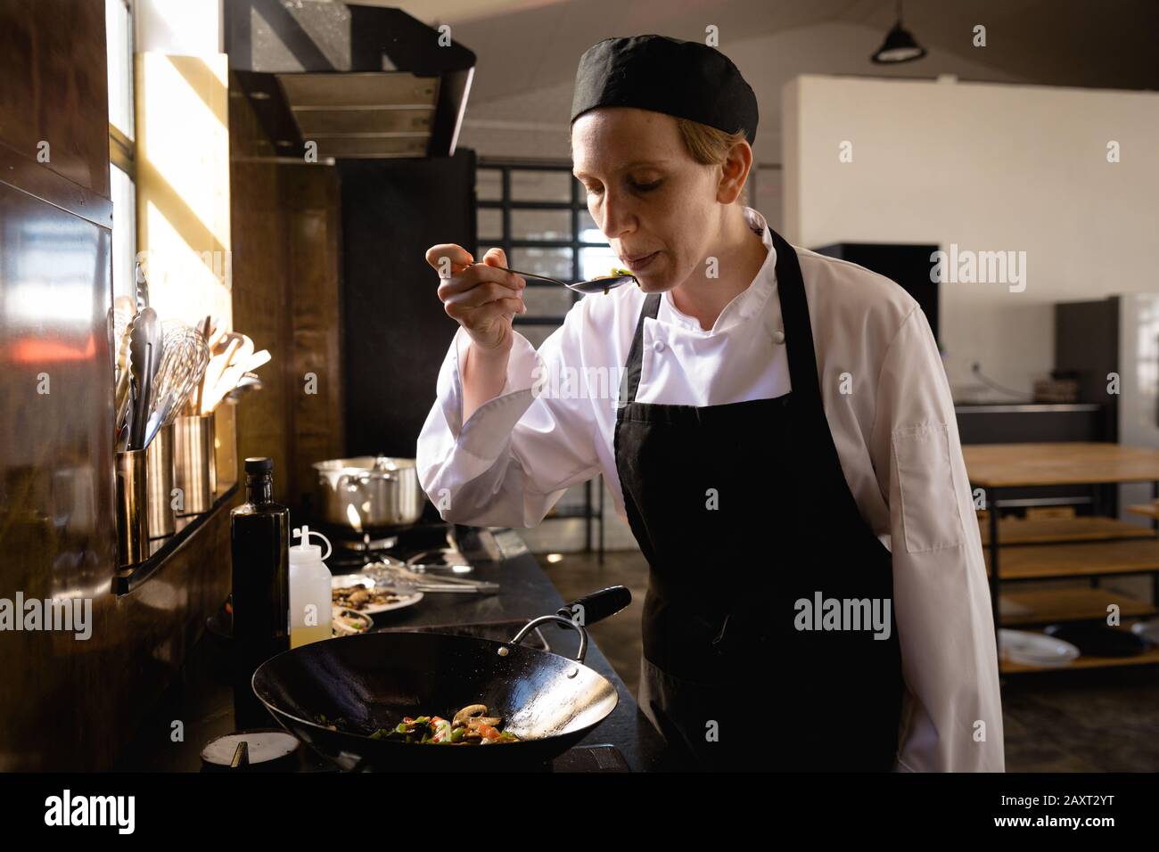 Vue de face d'une femme caucasienne à un cours de cuisine dans une cuisine de restaurant, soufflant des légumes chauds sur une cuillère avant de les goûter. Enjo des aînés actifs Banque D'Images