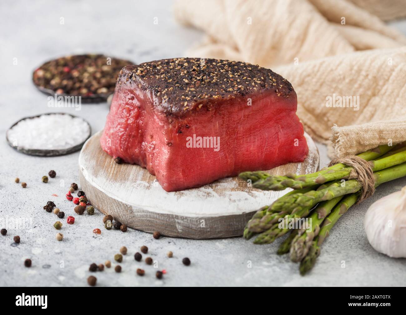 Tranche de bœuf cru joint de topaside avec sel et poivre sur planche à découper ronde avec pointes d'asperges et ail sur fond de table de cuisine léger. Banque D'Images