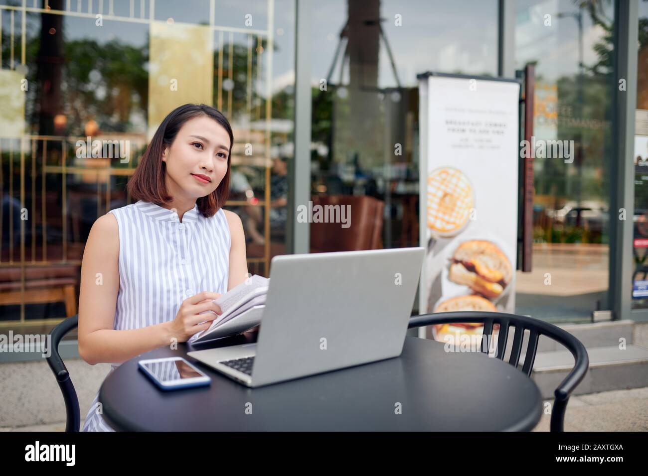 Belle confiance woman in casual clothing reading book et de boire du café tout en restant assis dans un café avec un ordinateur portable et ordinateur portable Banque D'Images