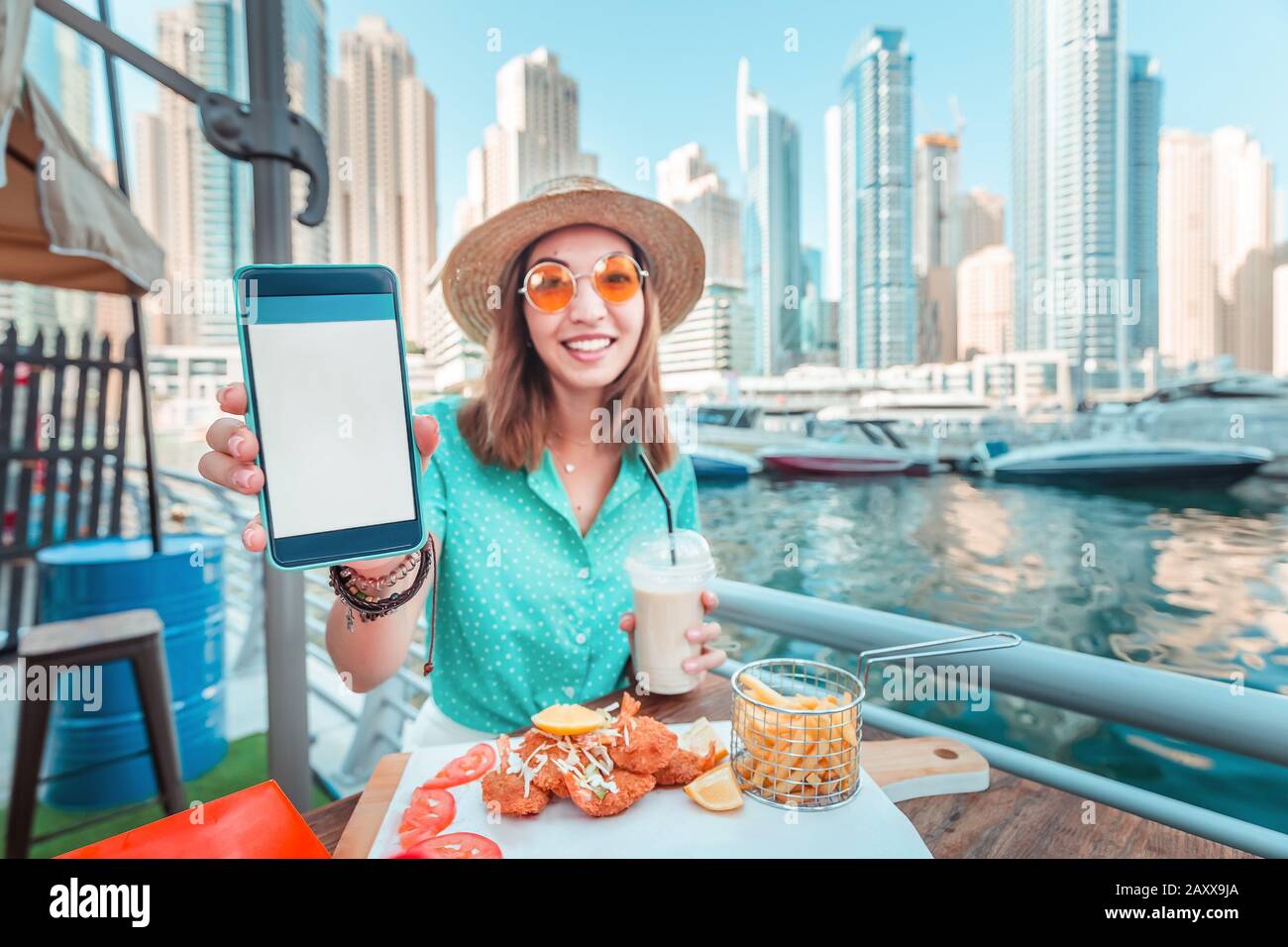 La jeune fille asiatique mange de la cuisine de fruits de mer surplombant les gratte-ciel et le port de plaisance de Dubaï et montre un écran vide de son smartphone. Concept d'application pour CA Banque D'Images