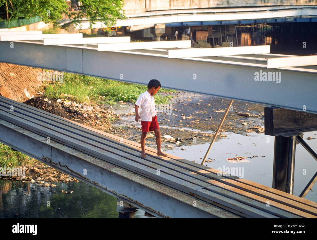 Un jeune garçon marche le long de tuyaux traversant un canal parsemé de détritus, Jakarta, Indonésie, juin 1995 Banque D'Images