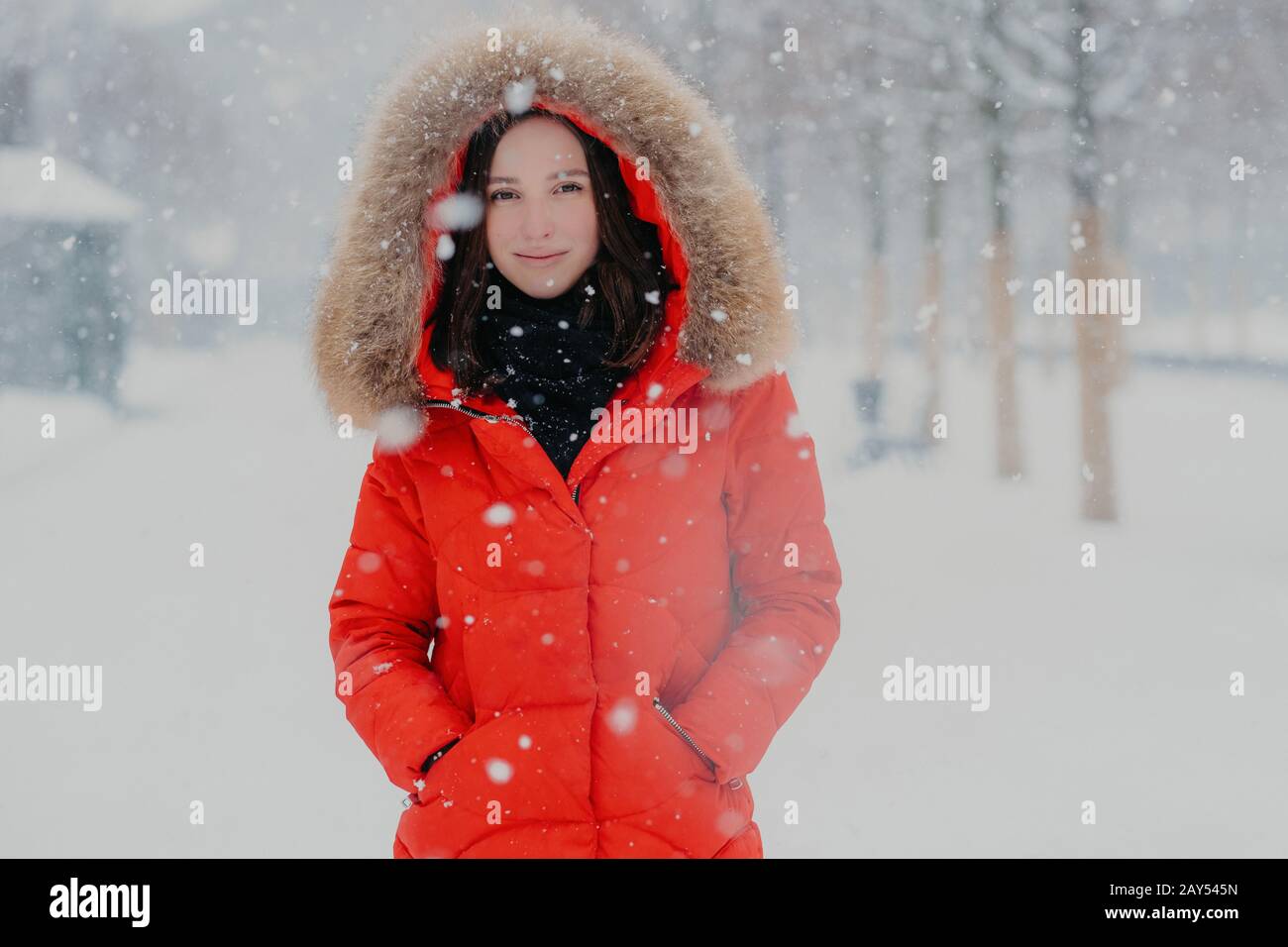 Une femme souriante et attrayante avec une peau saine, des cheveux sombres, porte une veste rouge, maintient les mains dans les poches par temps froid, résiste Banque D'Images