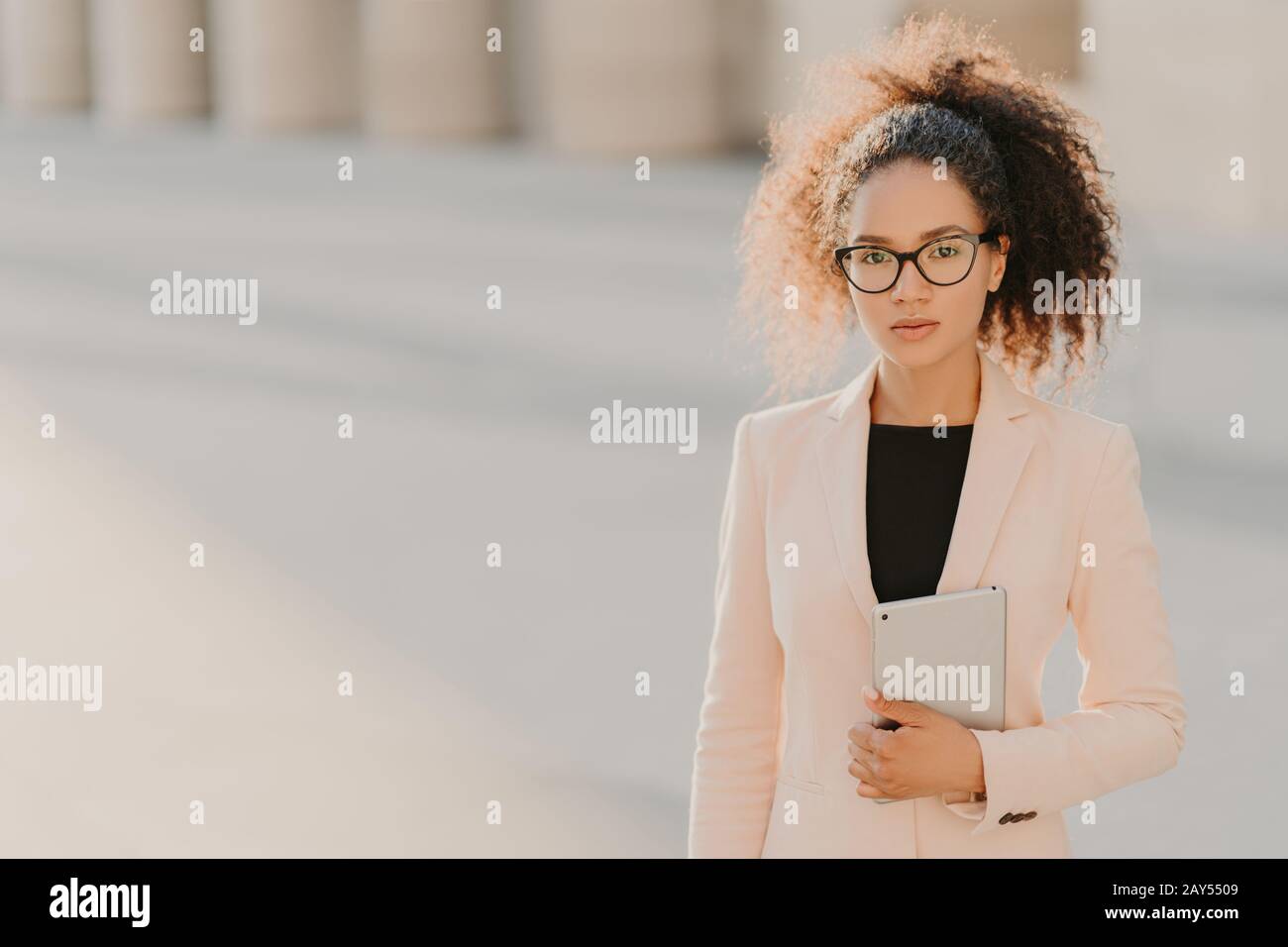 Photo de l'élégante femme d'affaires afro-américaine utilise une tablette d'ordinateur extérieur, porte une veste blanche formelle, lunettes transparentes avec cadres noirs, curieusement Banque D'Images