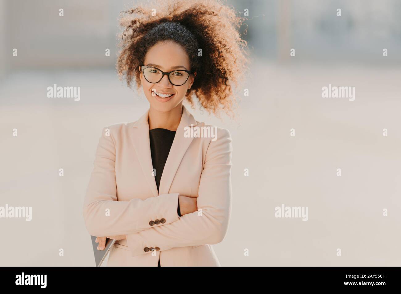 Une femme d'affaires joyeuse et confiante avec ses cheveux rafles d'Afro, maintient les bras pliés, utilise un ordinateur tablette moderne, sourit positivement, attend le travail par Banque D'Images