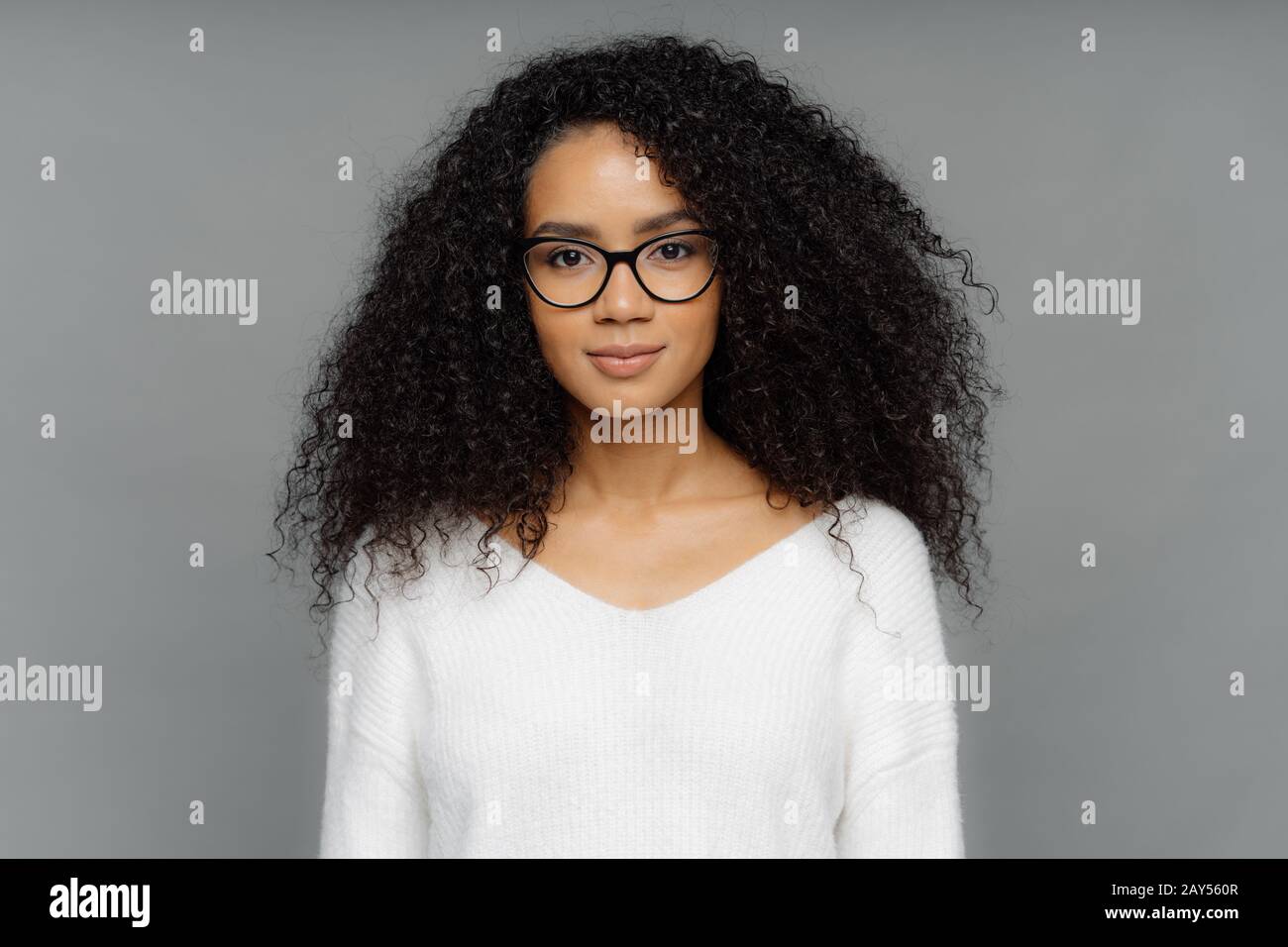 Portrait d'une femme sérieuse avec peau foncée, Afro bushy cheveux, porte de grands verres transparents et blanc doux chandail, regarde directement à l'appareil photo, isolé ov Banque D'Images