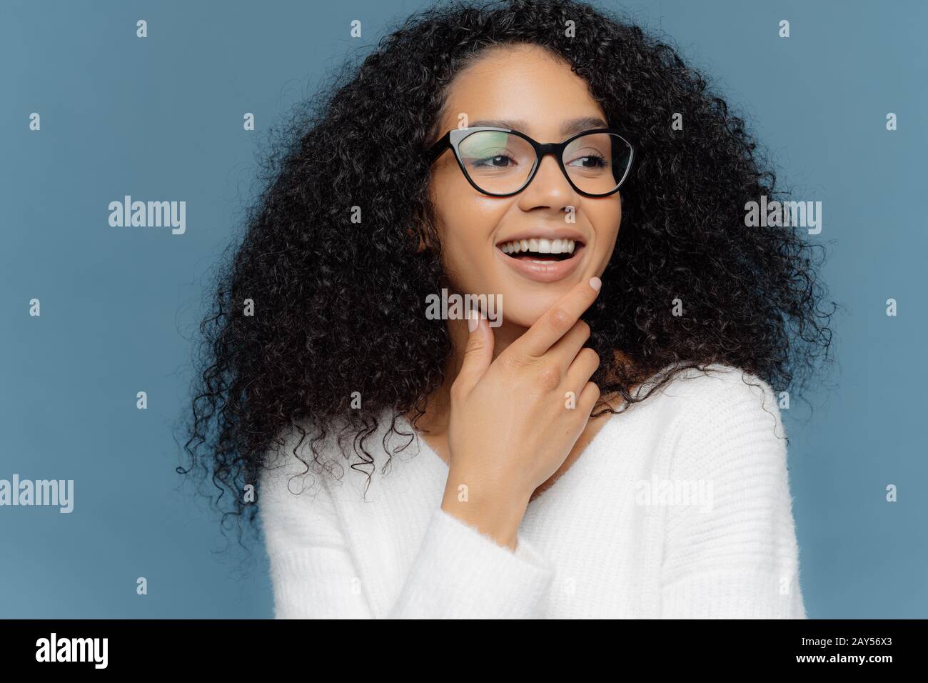 Très gros plan de la peau foncée agréable à la jeune femme est titulaire d'chin, regarde ailleurs, a les cheveux bouclés, large sourire, être en bonne humeur, isolé sur zone bleue Banque D'Images