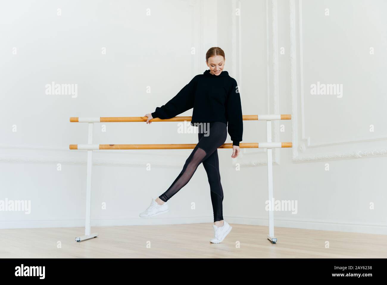 Jeune danseuse de ballet se réchauffe, se tient près des mains courantes, vêtu de vêtements de sport noirs, se tient sur la pointe de la chaussure, porte des sneakers blanches, focalisées sur le sol, Banque D'Images