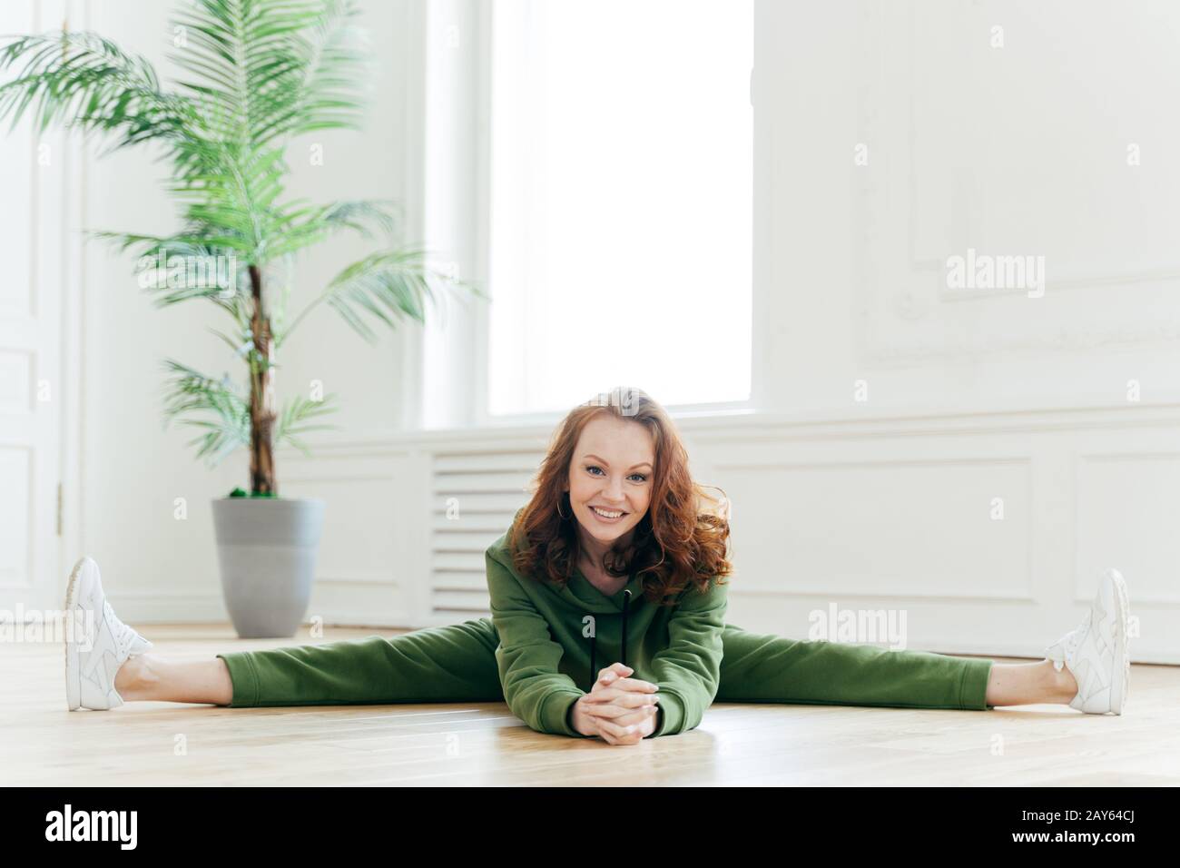 Femme mince et flexible avec des cheveux de gingembre, se décompose la jambe, a des exercices d'étirements, pose à l'intérieur de la maison, a le sourire doux sur le visage, porte vert tracksui Banque D'Images