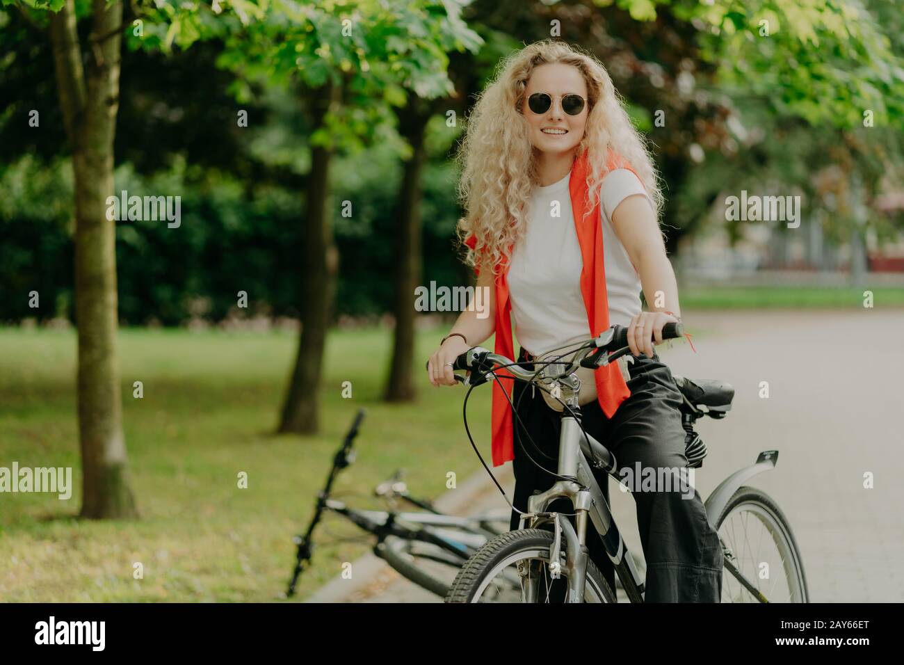Content actif curly haired femme vélo couvre la distance sur son propre transport, porte des lunettes de soleil, tenue décontractée, pose dans le parc avec des arbres verts, expre Banque D'Images