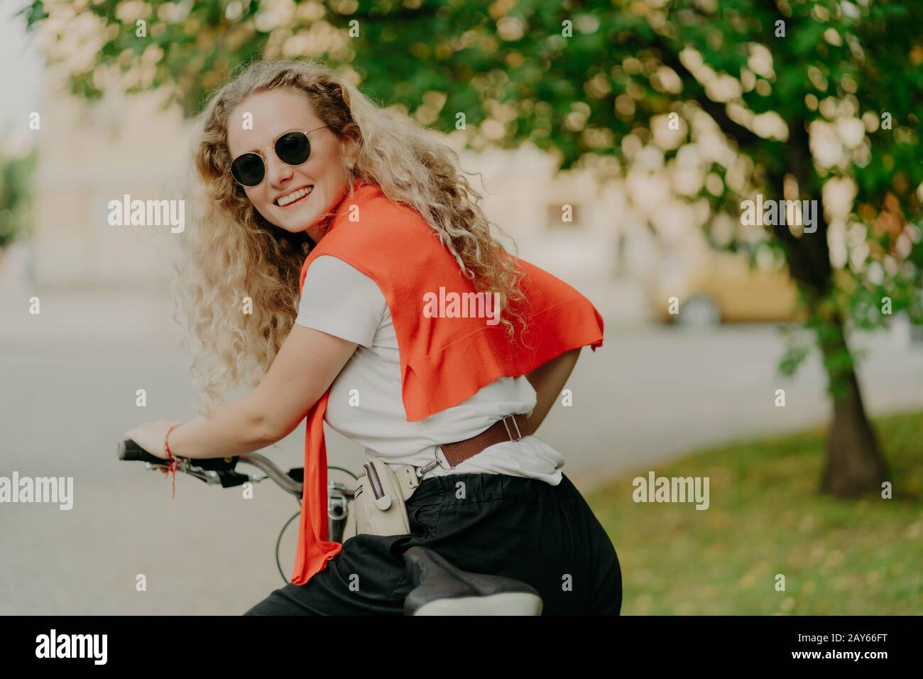 Photo de la femme enchantée pose de nouveau à l'appareil photo, fait des promenades à vélo à l'extérieur, porte des lunettes de soleil, t-shirt et sweat-shirt sur les épaules, pose près de l'arbre vert, a t Banque D'Images