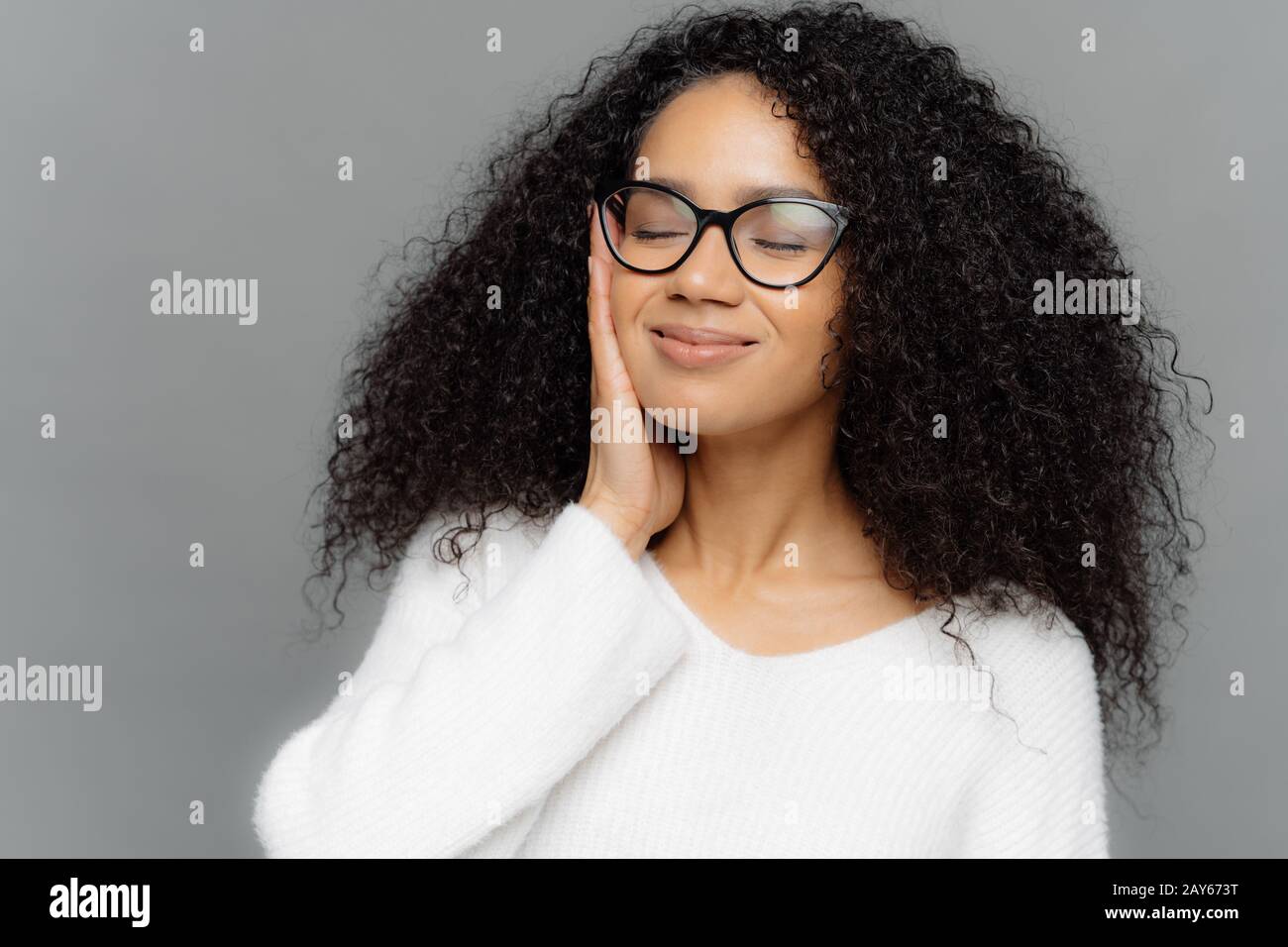 Portrait d'une femme à peau sombre et satsifiée avec des cheveux frisés, les yeux fermés, touche la joue avec les mains, imagine quelque chose d'agréable, porte des lunettes optiques Banque D'Images