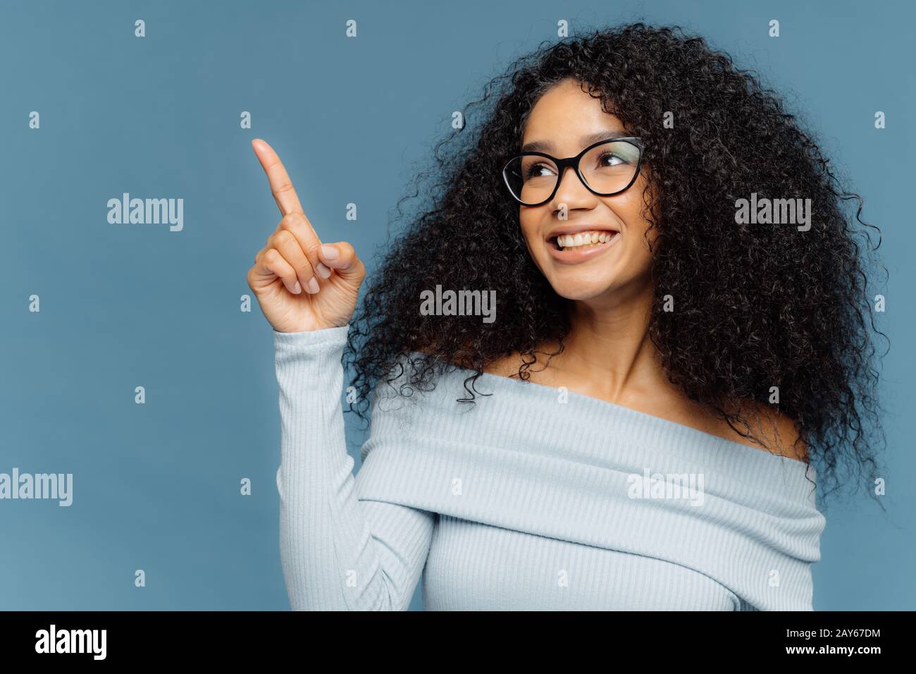 Une femme sourit et sourit avec une peau sombre et heureuse, avec des points de coiffure Afro, index finger vers le haut, montre quelque chose sur l'espace vide, a une expression heureuse, porte Banque D'Images