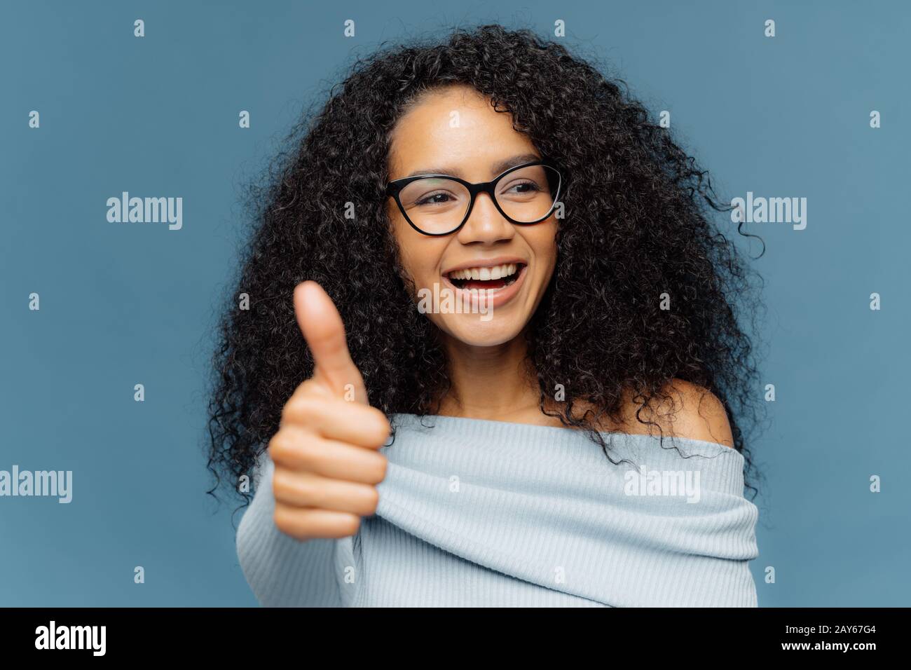 J'aime ça ! Portrait d'une femme heureuse avec Afro coiffure, montre le  pouce vers le haut, donne une opinion positive, regarde de côté, porte des  lunettes optiques et sweat-shirt, isolat Photo Stock -