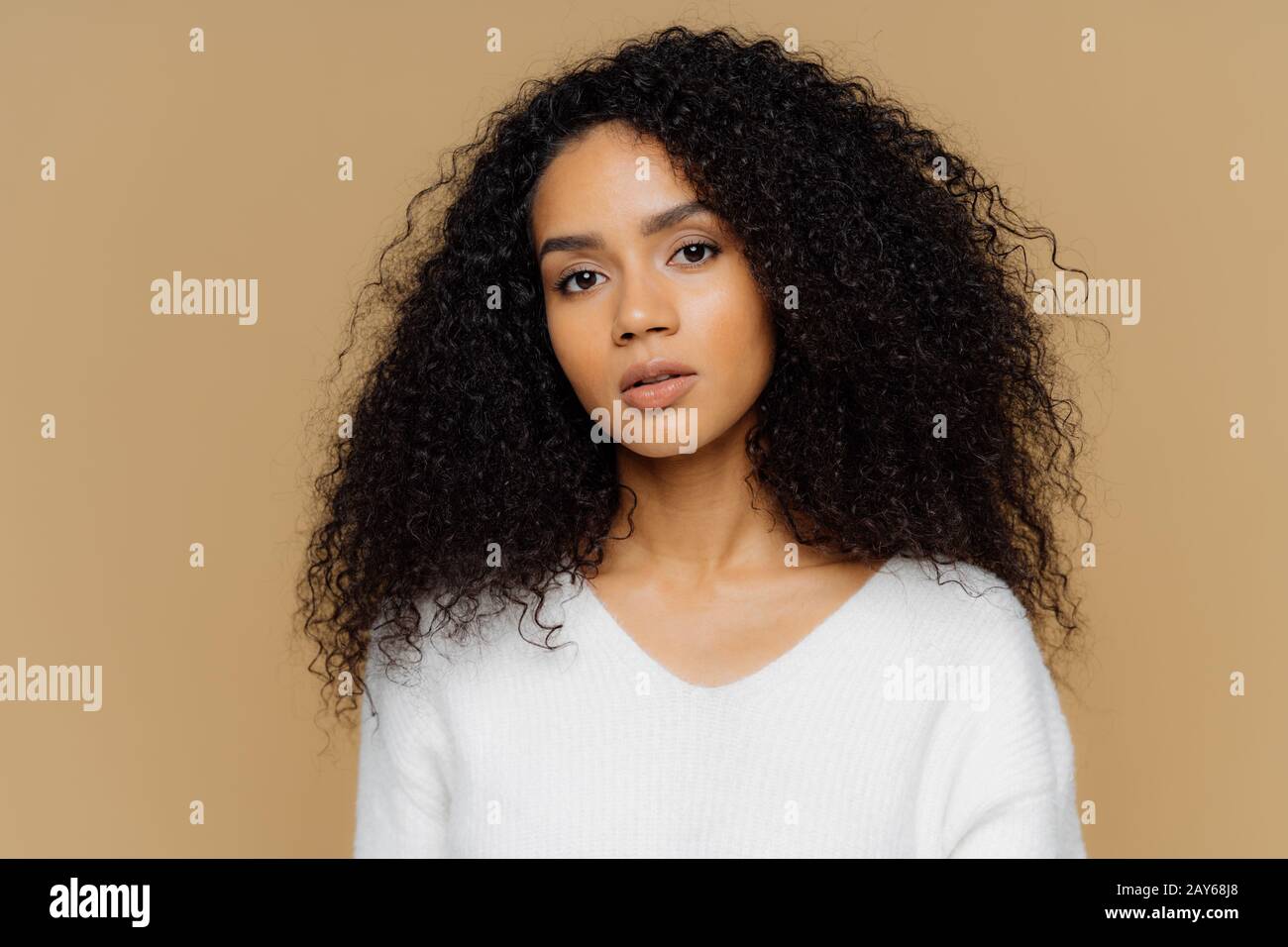 Portrait d'une belle femme sombre et sourenée avec des cheveux noirs frisés, a un maquillage minimal, regarde calmement l'appareil photo, porte un cavalier blanc, se tient à nouveau Banque D'Images