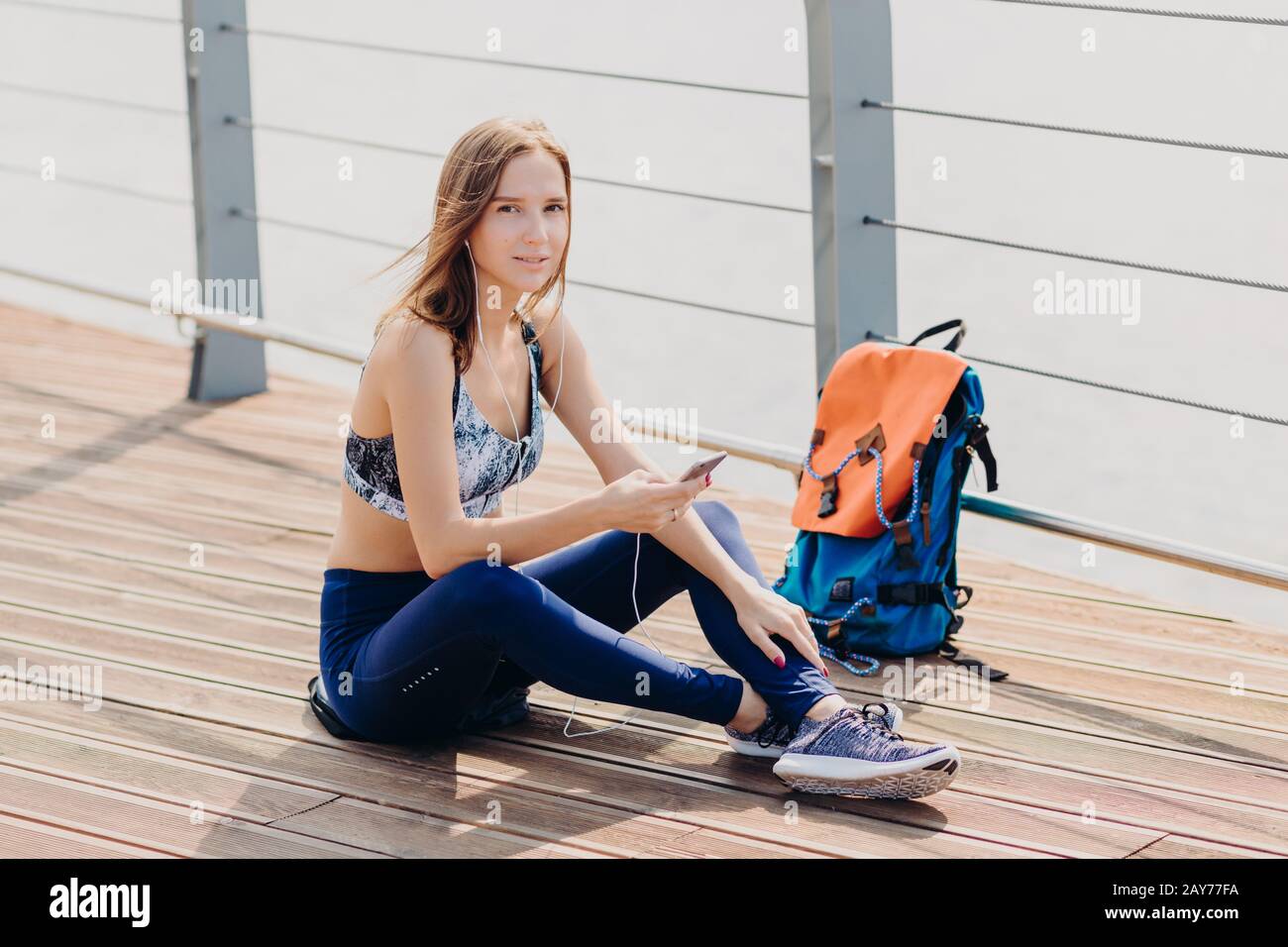 Jolie femme détendue avec des cheveux sombres, une peau pure saine, vêtue de vêtements de sport, tient le téléphone portable pour la communication, écoute la piste préférée, sac à dos s Banque D'Images