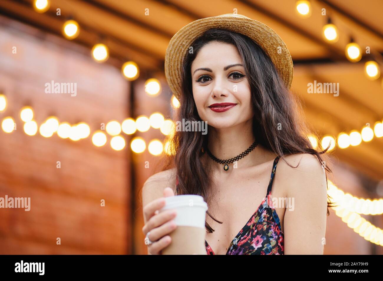 Portrait d'une dame brunette aux yeux charmants, aux lèvres rouges bien formées et à la peau pure, vêtue d'un chapeau et d'une chemise d'été, tenant du café frais à emporter en pa Banque D'Images