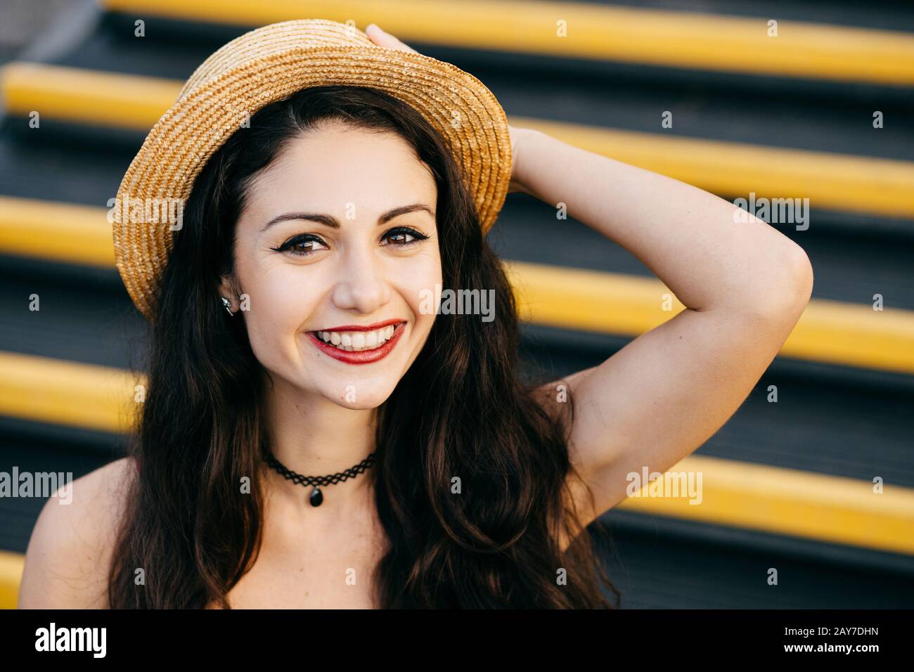 Une femme gaie avec des cheveux sombres, une peau saine, des yeux lumineux portant un chapeau de paille sur la tête assise dans les escaliers, en gardant la main sur la tête souriante largement au niveau du caméer Banque D'Images