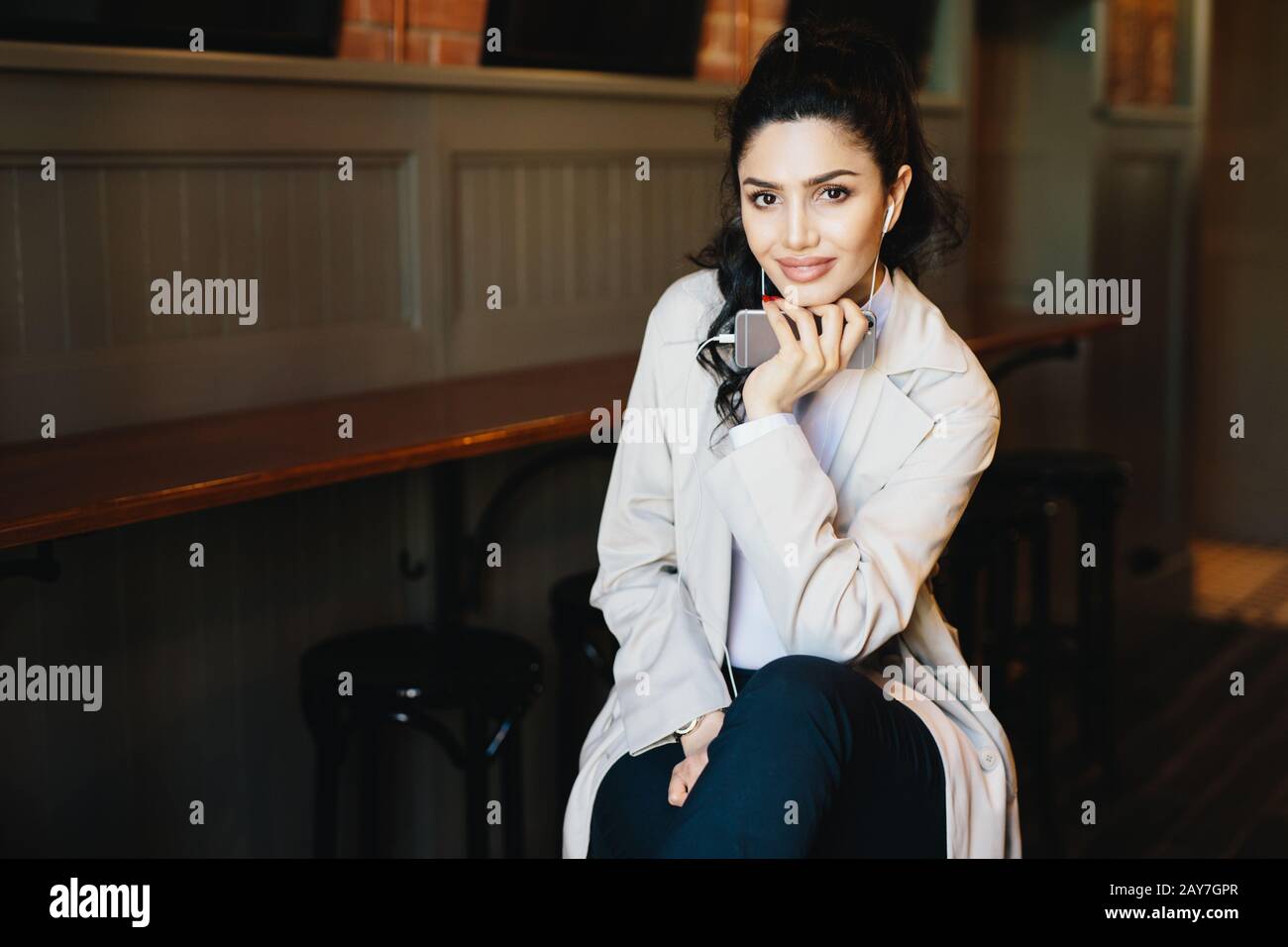 Portrait horizontal d'une femme à la mode avec des yeux sombres et des cheveux ayant une peau pure regardant dans l'appareil photo assis dans un café en profitant de la bonne musique pendant listenin Banque D'Images