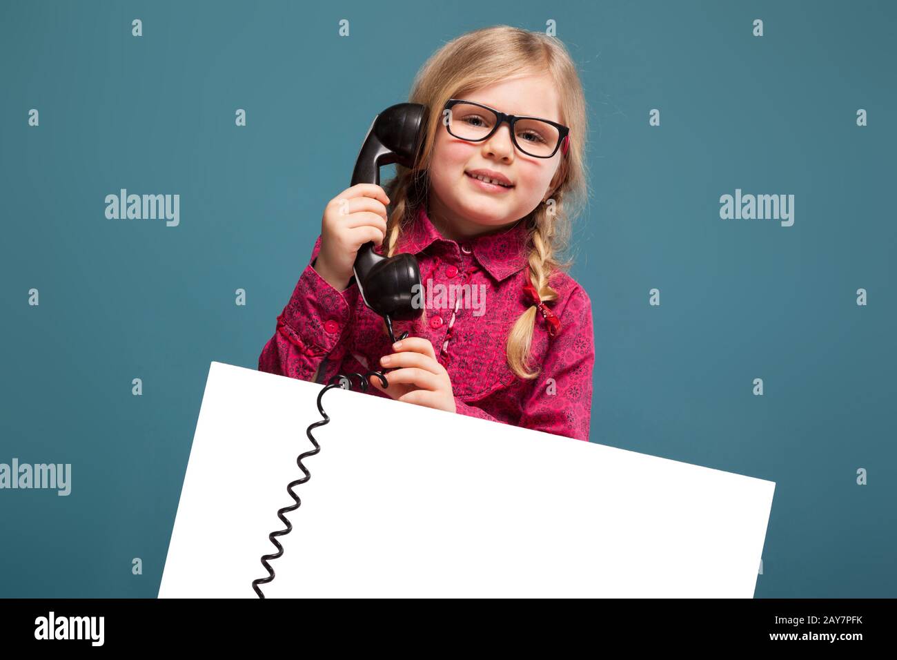 Jolie petite fille en chemise rose, pantalon et lunettes noirs tient une affiche vide et parle au téléphone Banque D'Images