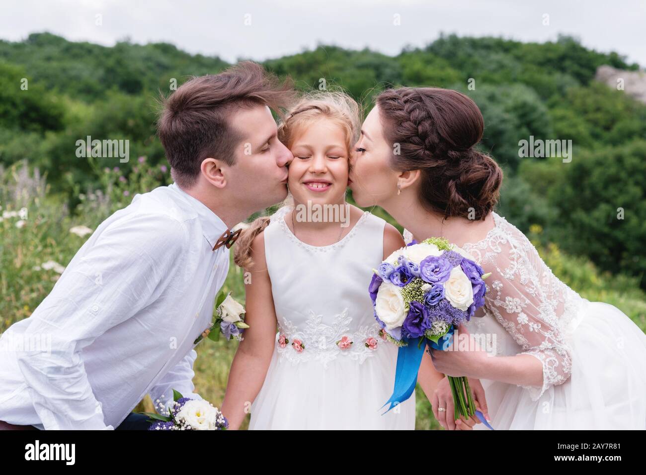 Les jeunes parents dans les robes de mariage embrassent leur jeune fille dans les joues Banque D'Images