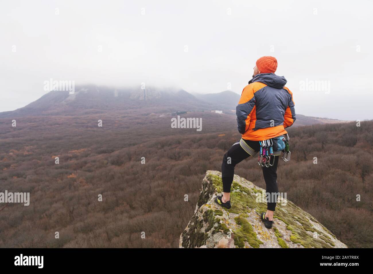 Hipster - un grimpeur dans une veste en duvet et un pied à casquette tricoté et repose sur le sommet d'une roche Banque D'Images
