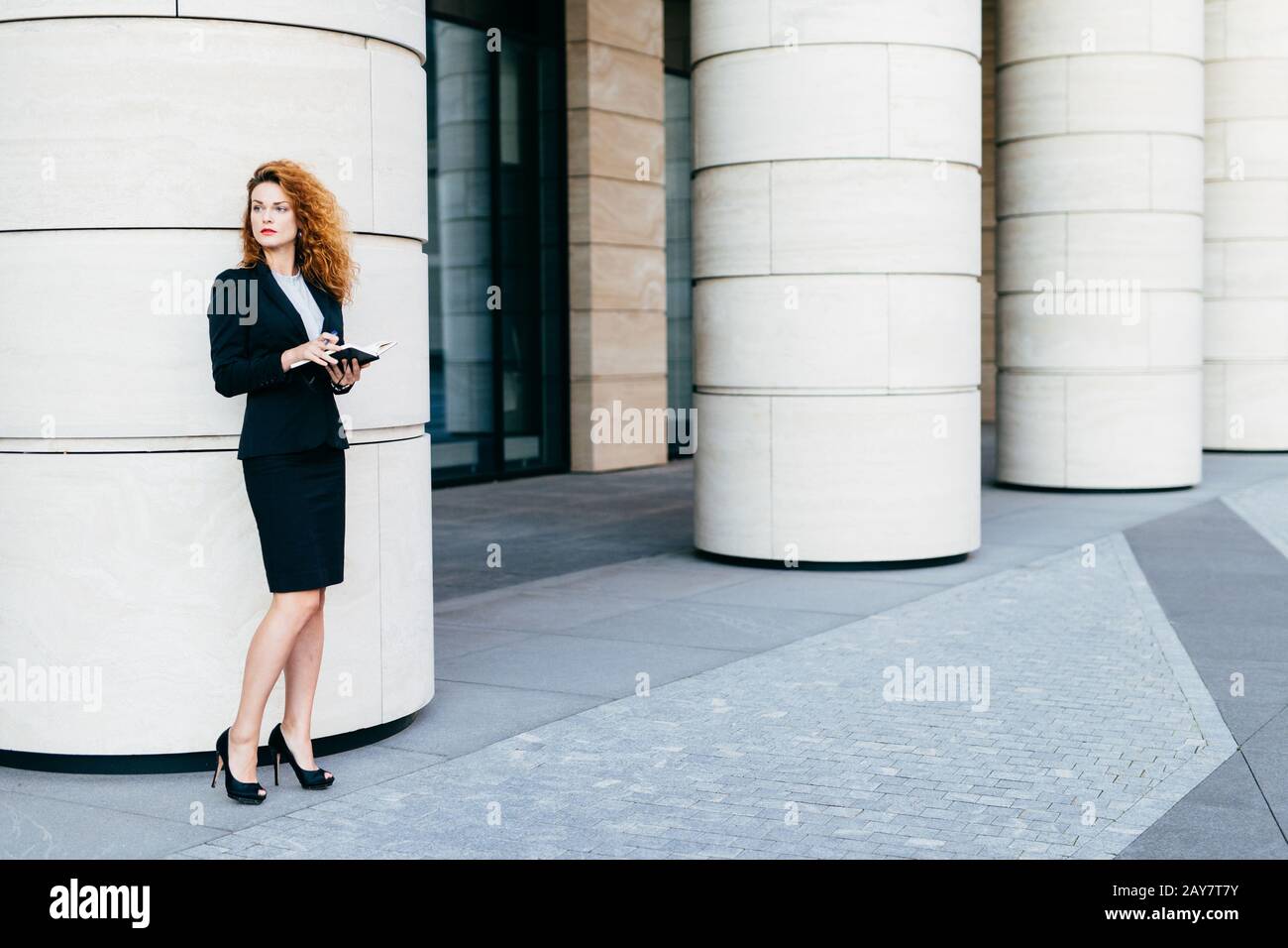 Portrait horizontal d'une femme d'affaires de bonne qualité vêtue de  vêtements habillés et de chaussures noires avec talons hauts, tenant un  livre de poche, en se tenant à côté de t Photo