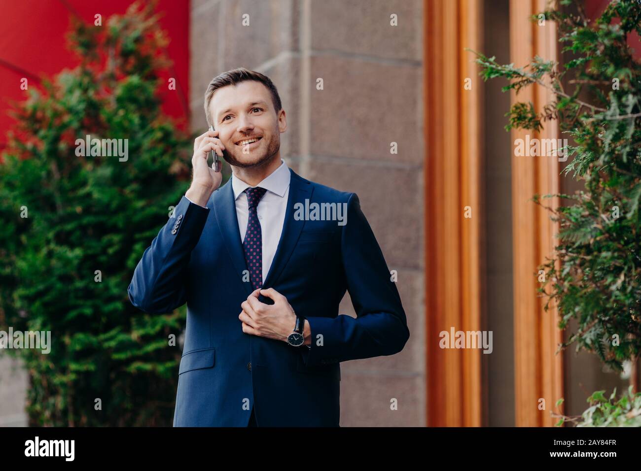 Portrait de beau homme d'affaires satisfait avec succès des discussions sur le téléphone cellulaire, traite des transactions bancaires, vêtu d'un costume d'entreprise, a une exp positive Banque D'Images