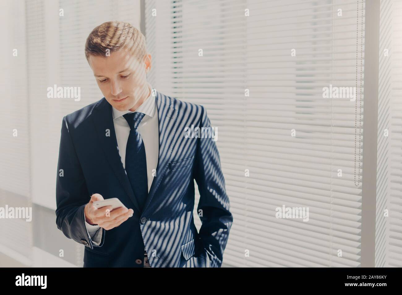 Jeune entrepreneur réussi en costume cher, tient le téléphone mobile, se tient au bureau, contre la fenêtre dans le cabinet, chats en ligne, lit des nouvelles sur finan Banque D'Images